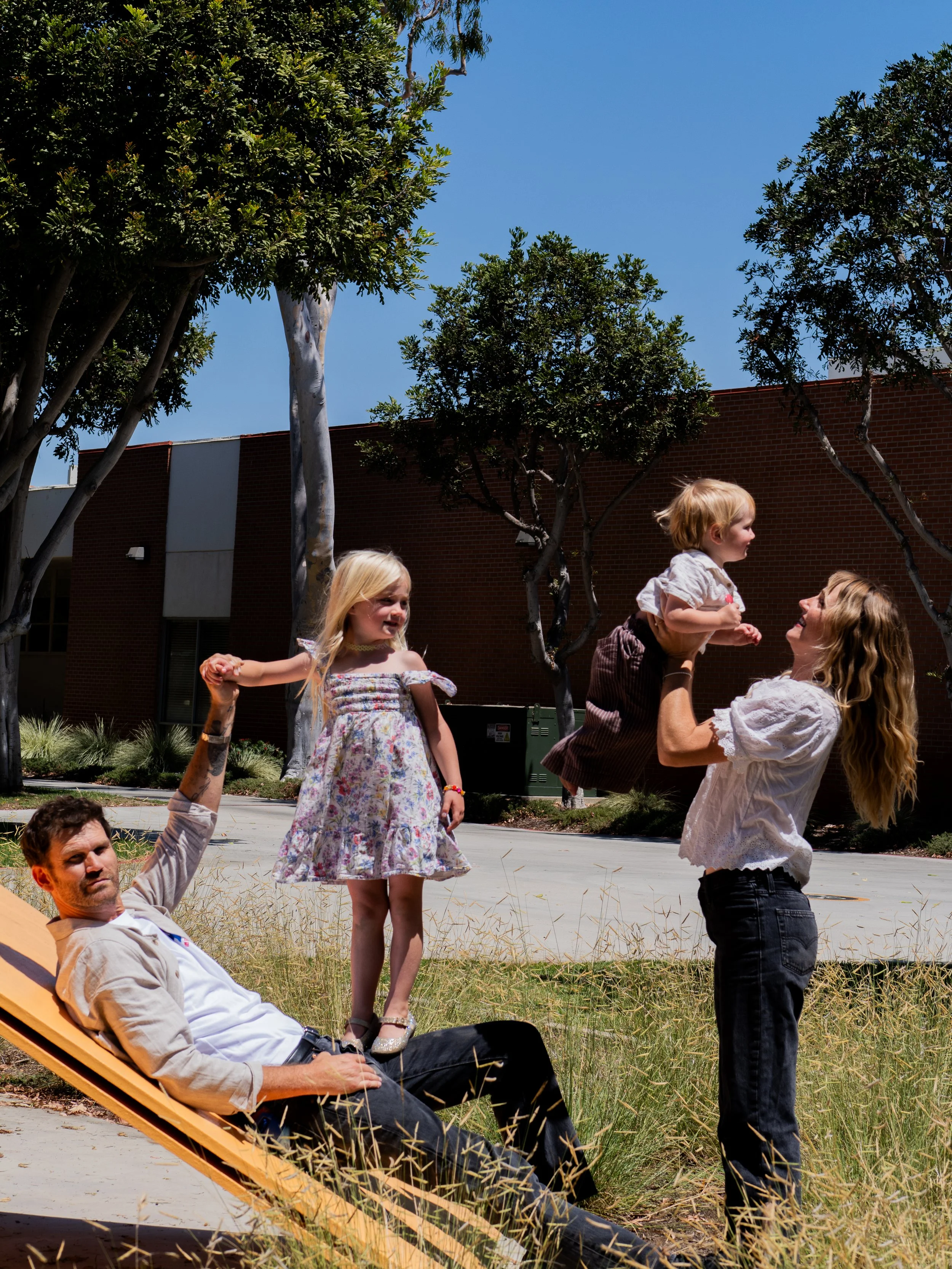 A group of people, including children and adults, are outdoors enjoying a sunny day. Some are standing and talking, while one person is sitting on a lounge chair. There are trees, grass, and a brick building in the background.