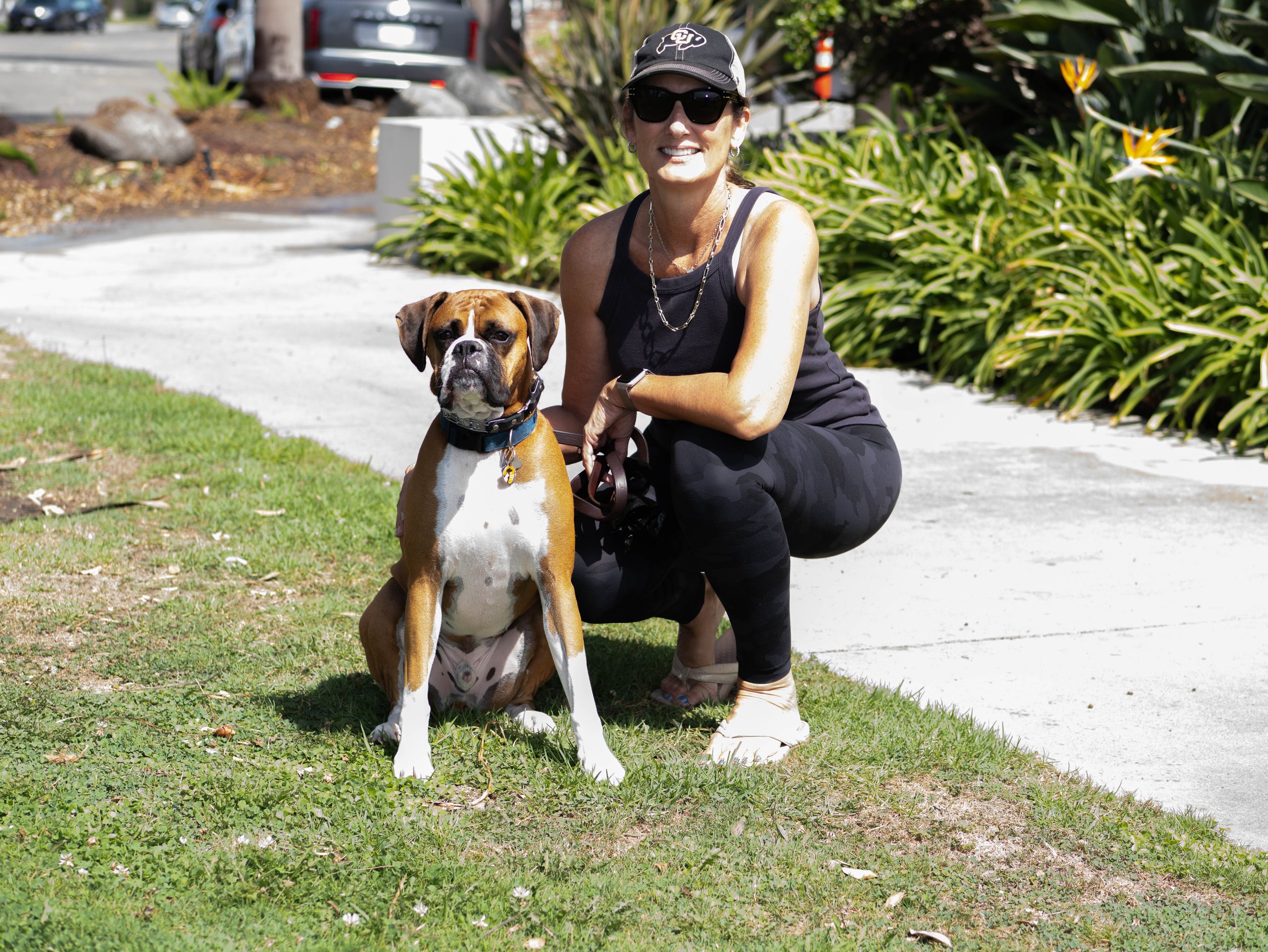 A woman in sunglasses, a baseball cap, and black athletic clothing kneels next to a brown and white Boxer dog on a grassy area, with greenery and parked cars in the background.