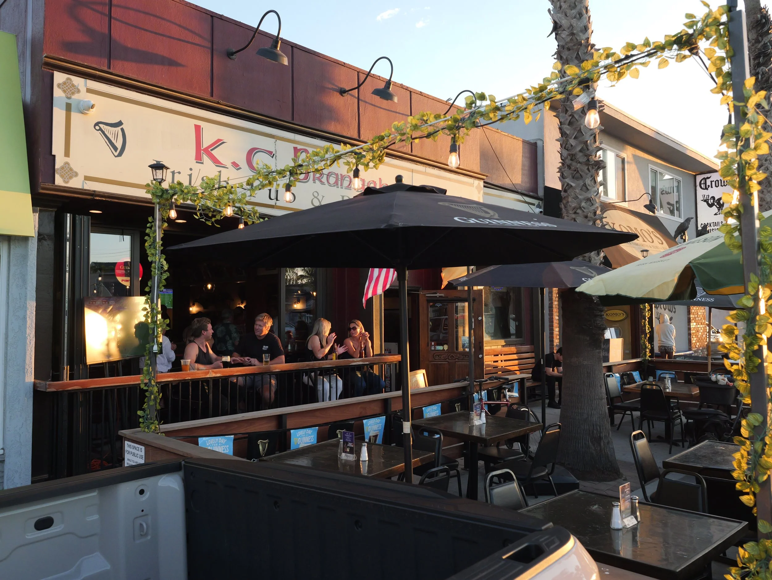 Outdoor patio with people sitting at tables under large umbrellas, string lights hanging above, palm trees, and a building with signs advertising Irish pub and cocktails.
