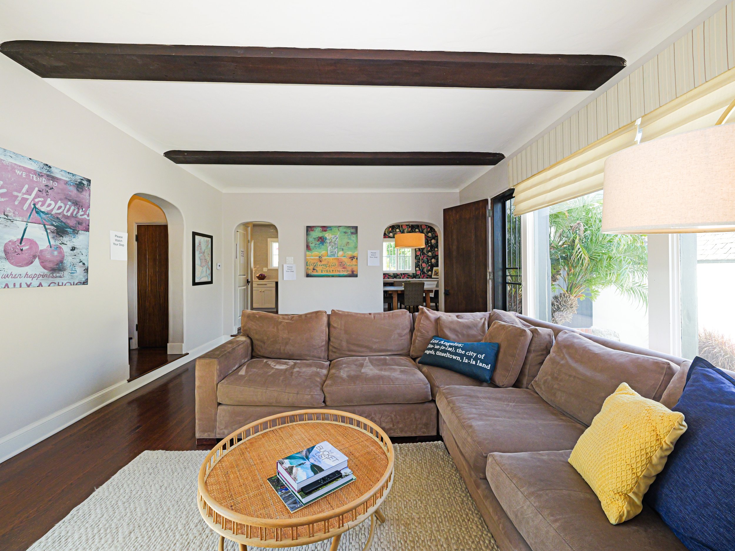 Living room with beige sectional sofa with yellow and navy pillows, round rattan coffee table with magazines, artwork on white walls, large window with blinds, wooden ceiling beams.