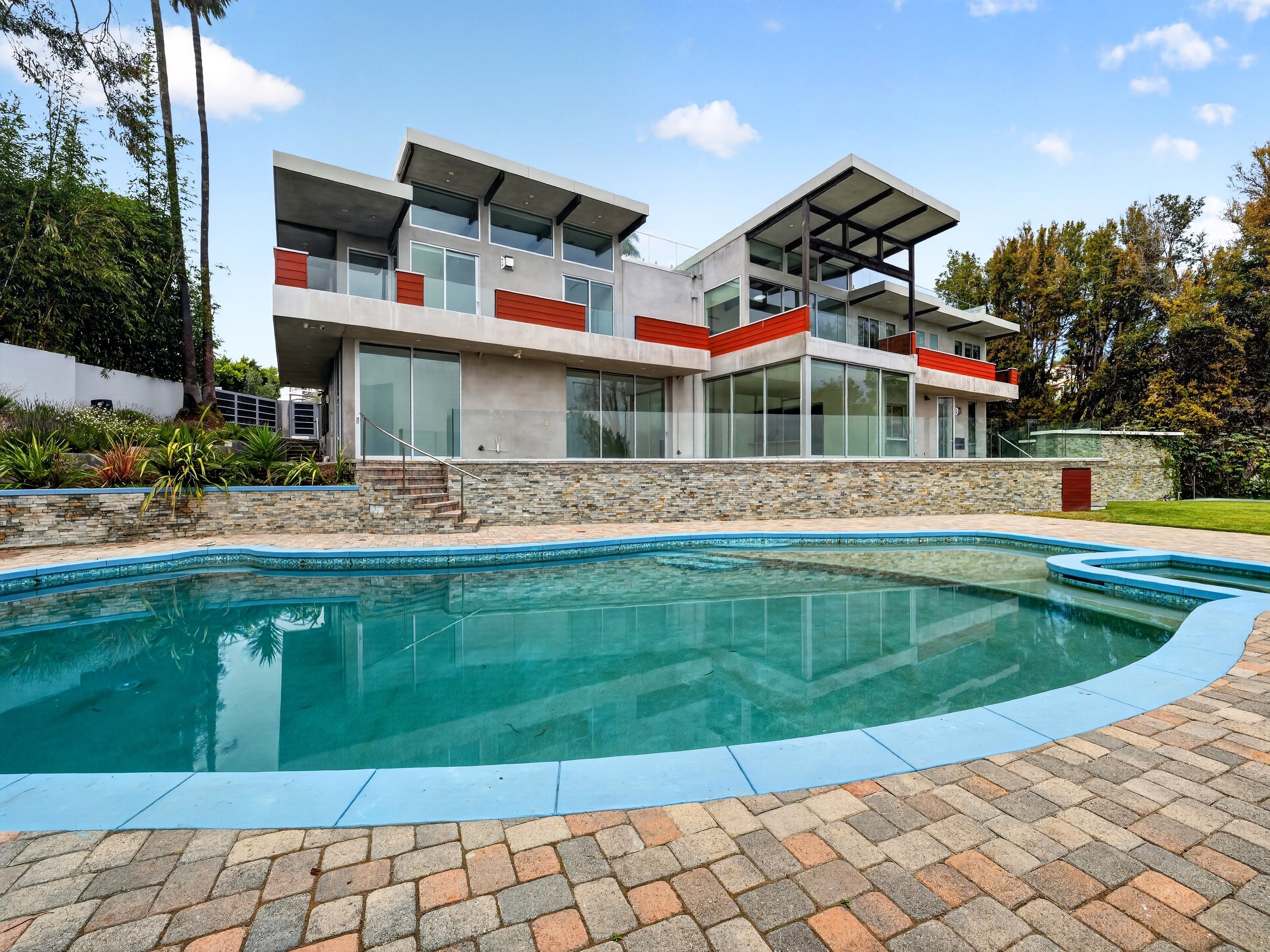Modern multi-story house with large glass windows and balconies, situated behind a swimming pool and surrounded by trees and landscaping.