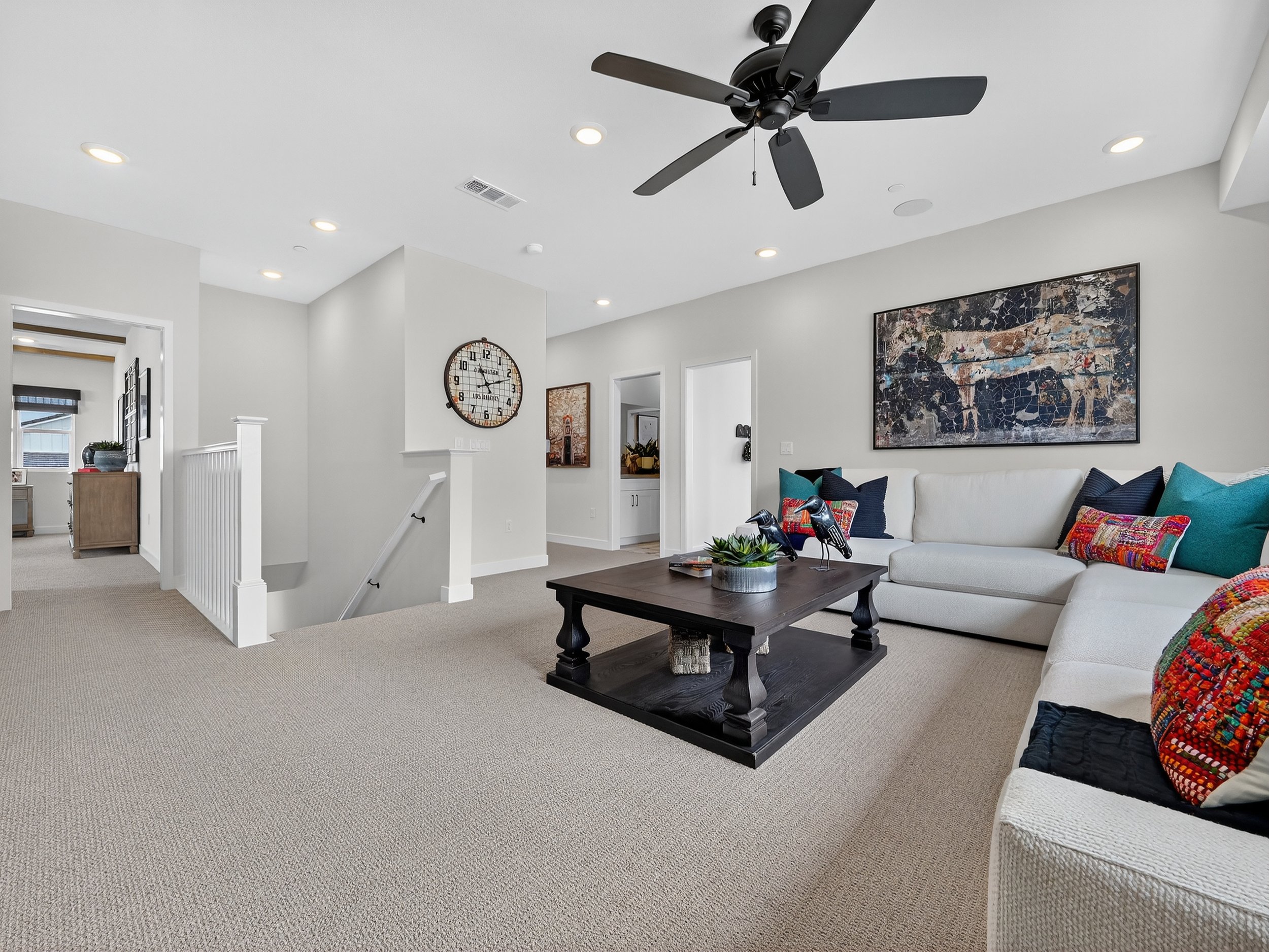 Living room with white walls, beige carpet, black ceiling fan, large wall clock, colorful pillows, and modern artwork.