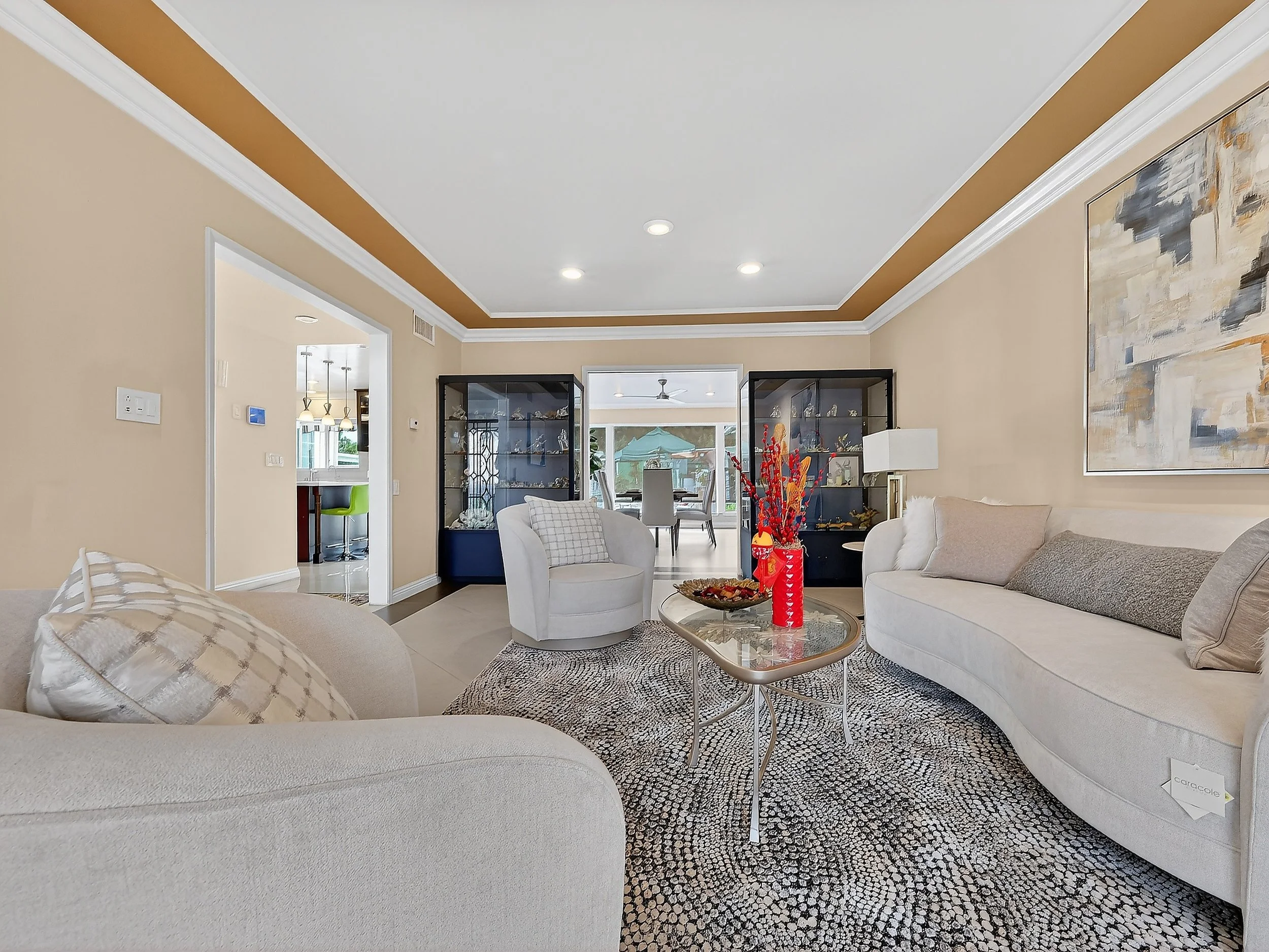 Living room with beige walls, white ceiling with brown border, and modern furniture including cream-tan sofas, a white armchair, and a glass coffee table with a red vase and decorative items. Black display cabinets and abstract artwork on walls, with sliding glass doors leading to an outdoor patio.