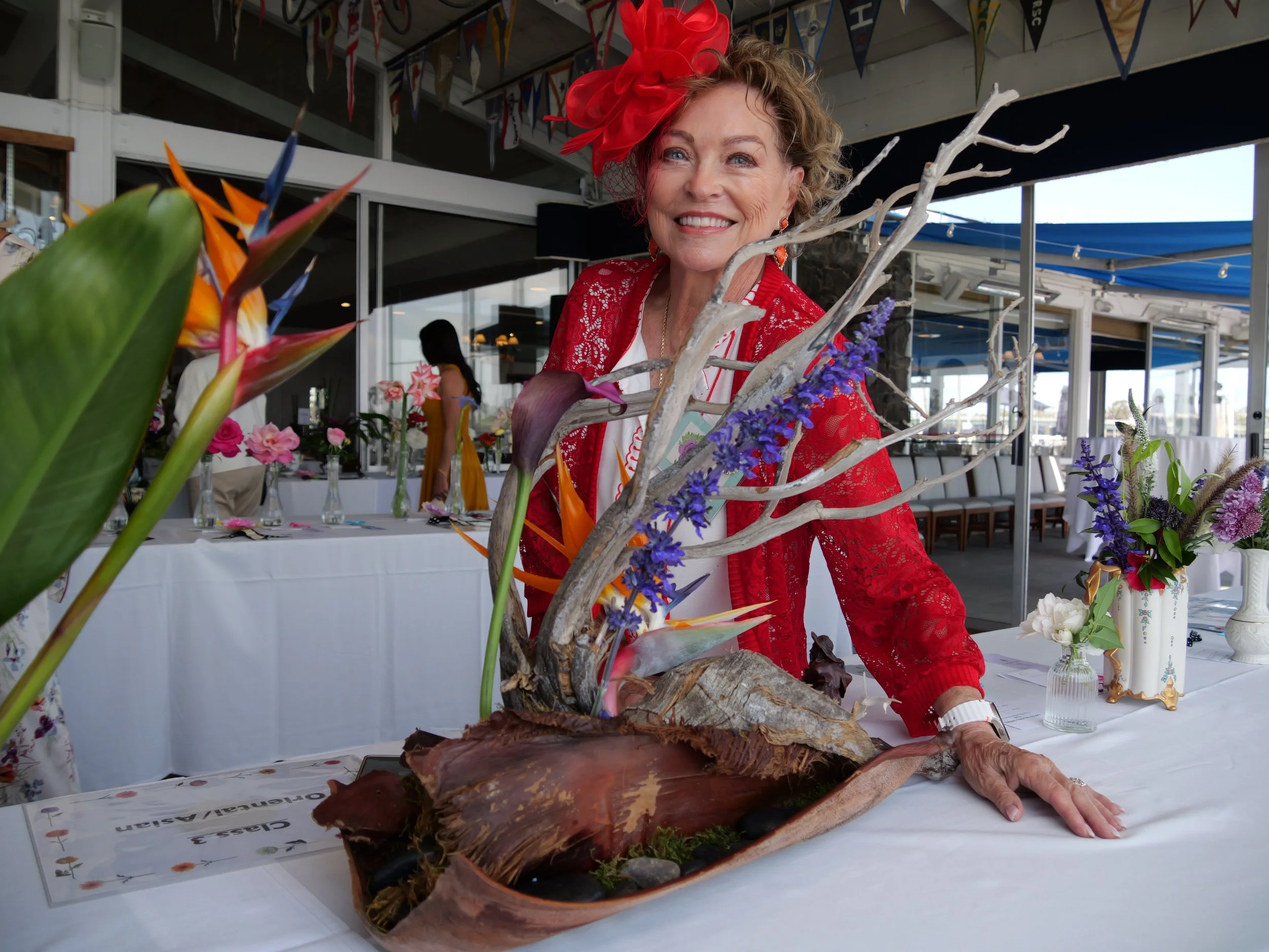 An elderly woman in red clothing and a red hat with a netting, smiling at a floral display at an indoor event. The display includes various flowers and a large piece of driftwood or wood sculpture. There are tables with flowers and decorations, and b