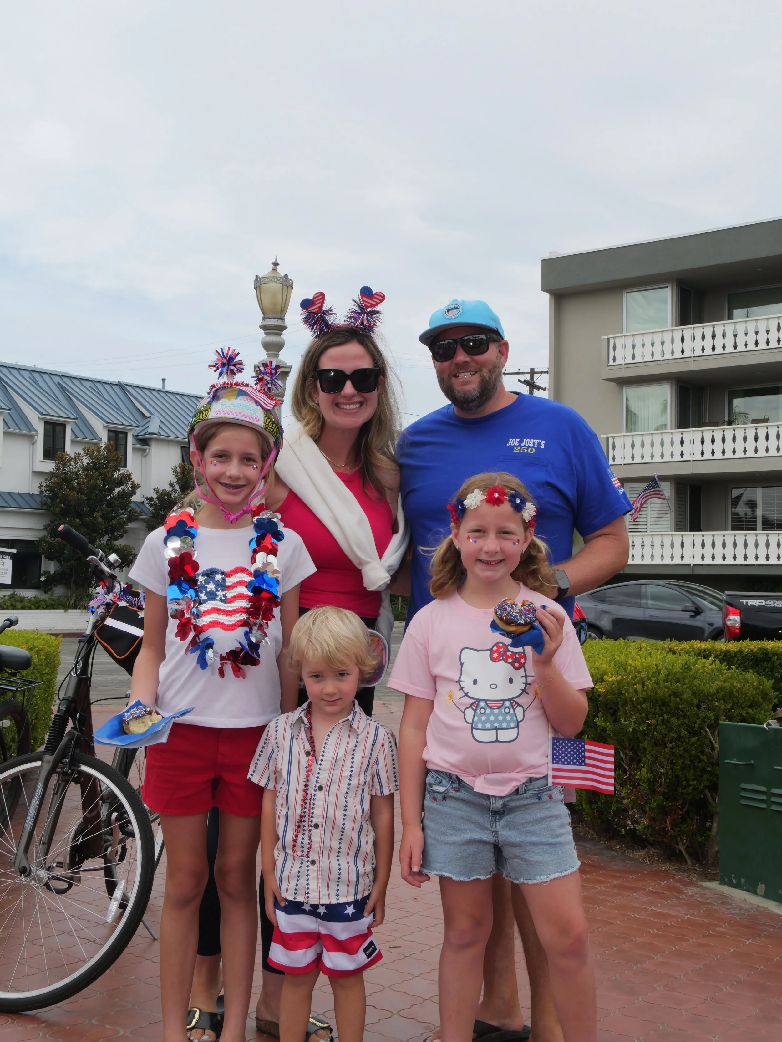 A group of people celebrating Independence Day with patriotic decorations, wearing red, white, and blue colors, and American flag accessories, on a city street with residential buildings in the background.