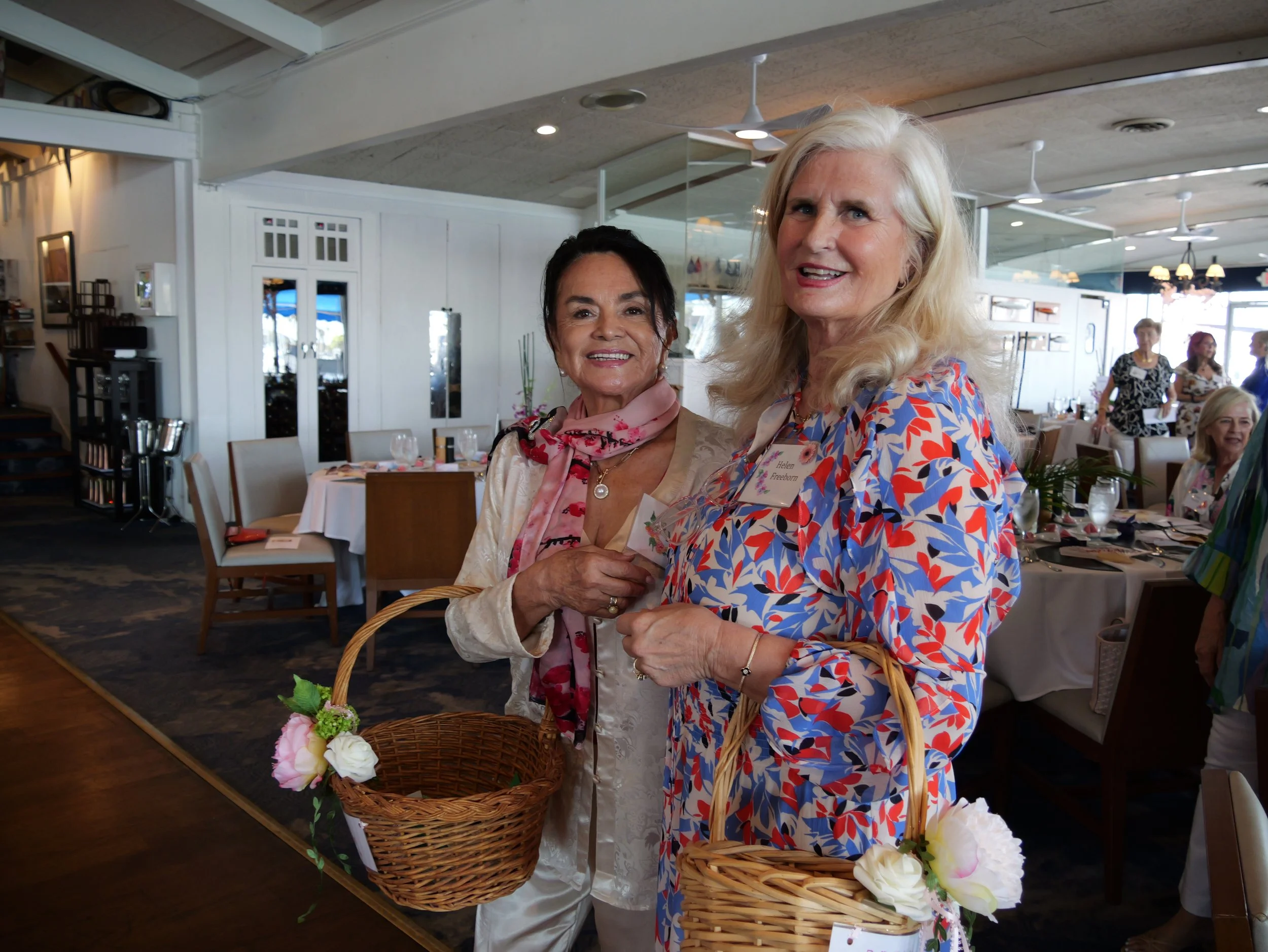 Two women standing inside a restaurant, holding baskets filled with flowers, smiling at the camera. The woman on the right is wearing a colorful floral dress and has blonde hair. The woman on the left is wearing a cream-colored outfit and a pink scar
