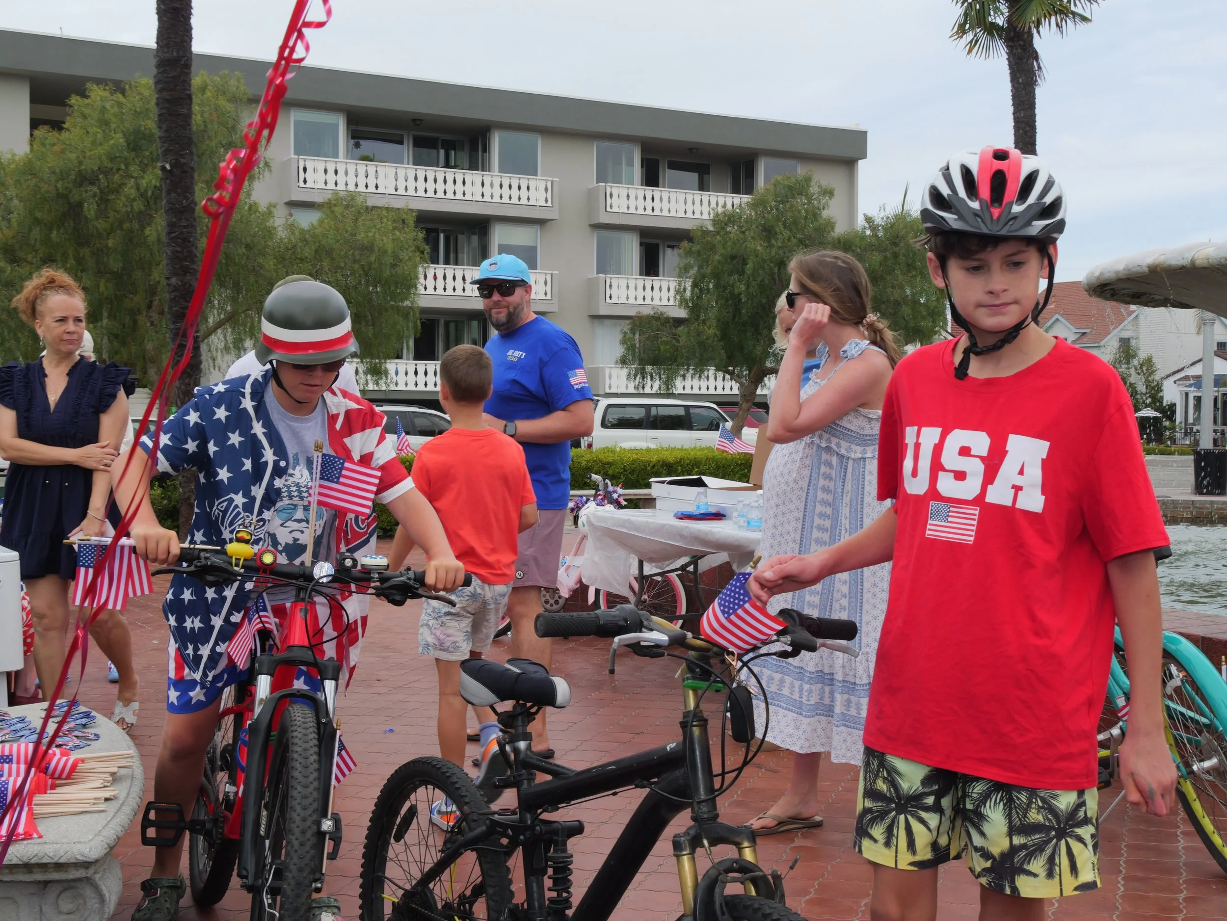 A group of people celebrating the Fourth of July with patriotic decorations, including American flags and themed clothing, outdoors near water with a building and trees in the background.