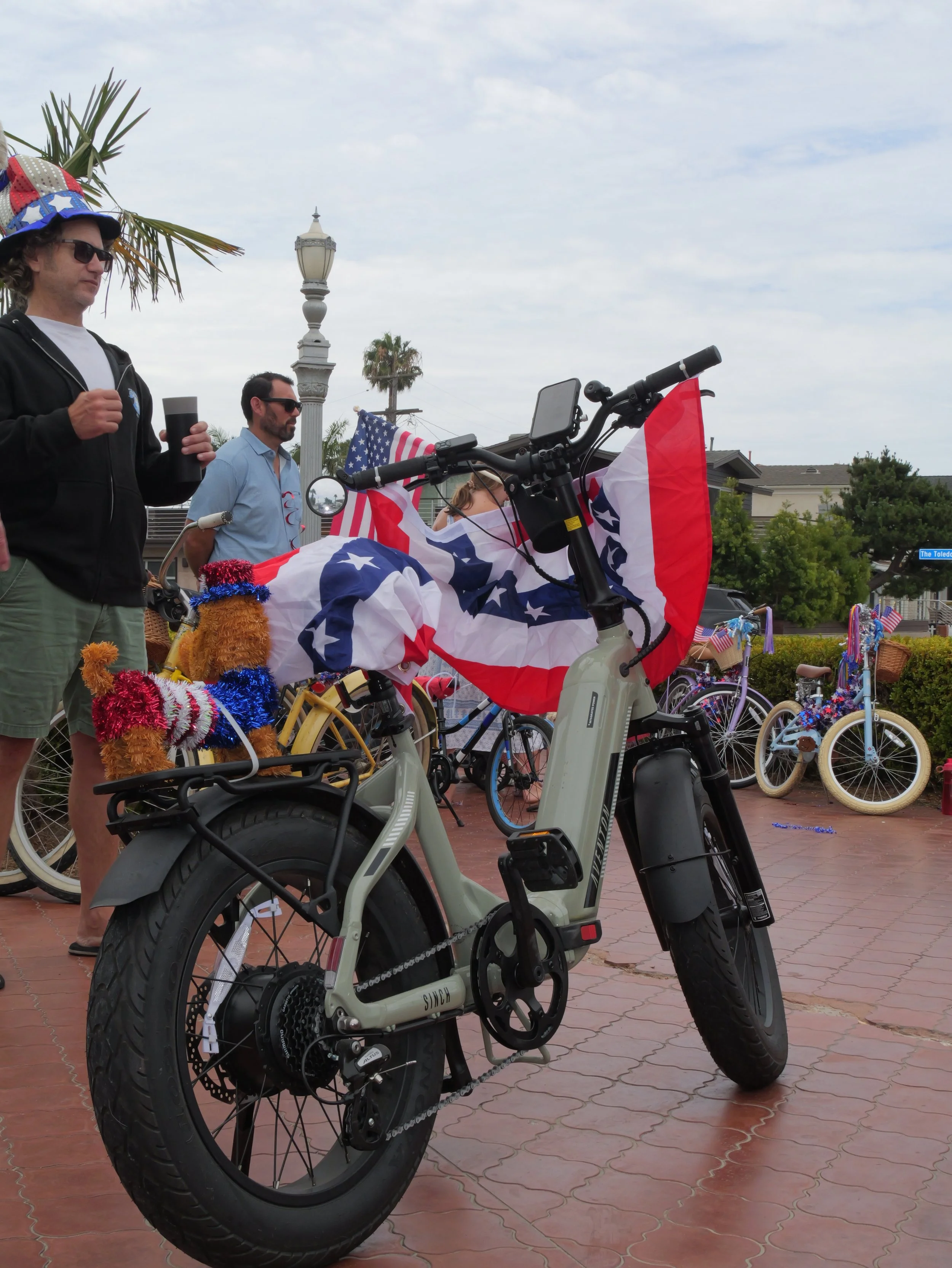 A group of people at a gathering with bicycles on display. The bicycles are decorated with American flags and patriotic decorations, and people are seen standing nearby, some wearing patriotic clothing and accessories. The scene appears to be outdoor