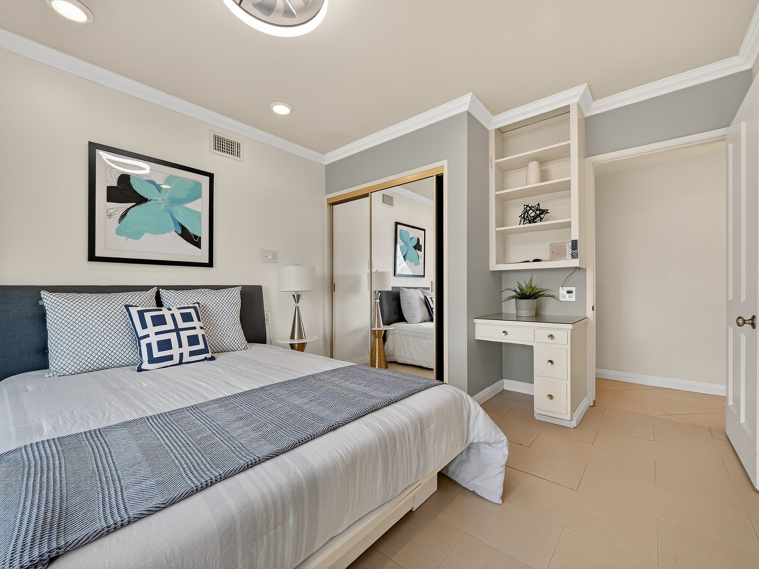 A bedroom with a bed, decorative pillows, two bedside lamps, and wall art featuring a blue butterfly. There is a built-in white shelf with a vase, a geometric sculpture, and a potted plant. A mirror and closet doors are also visible.