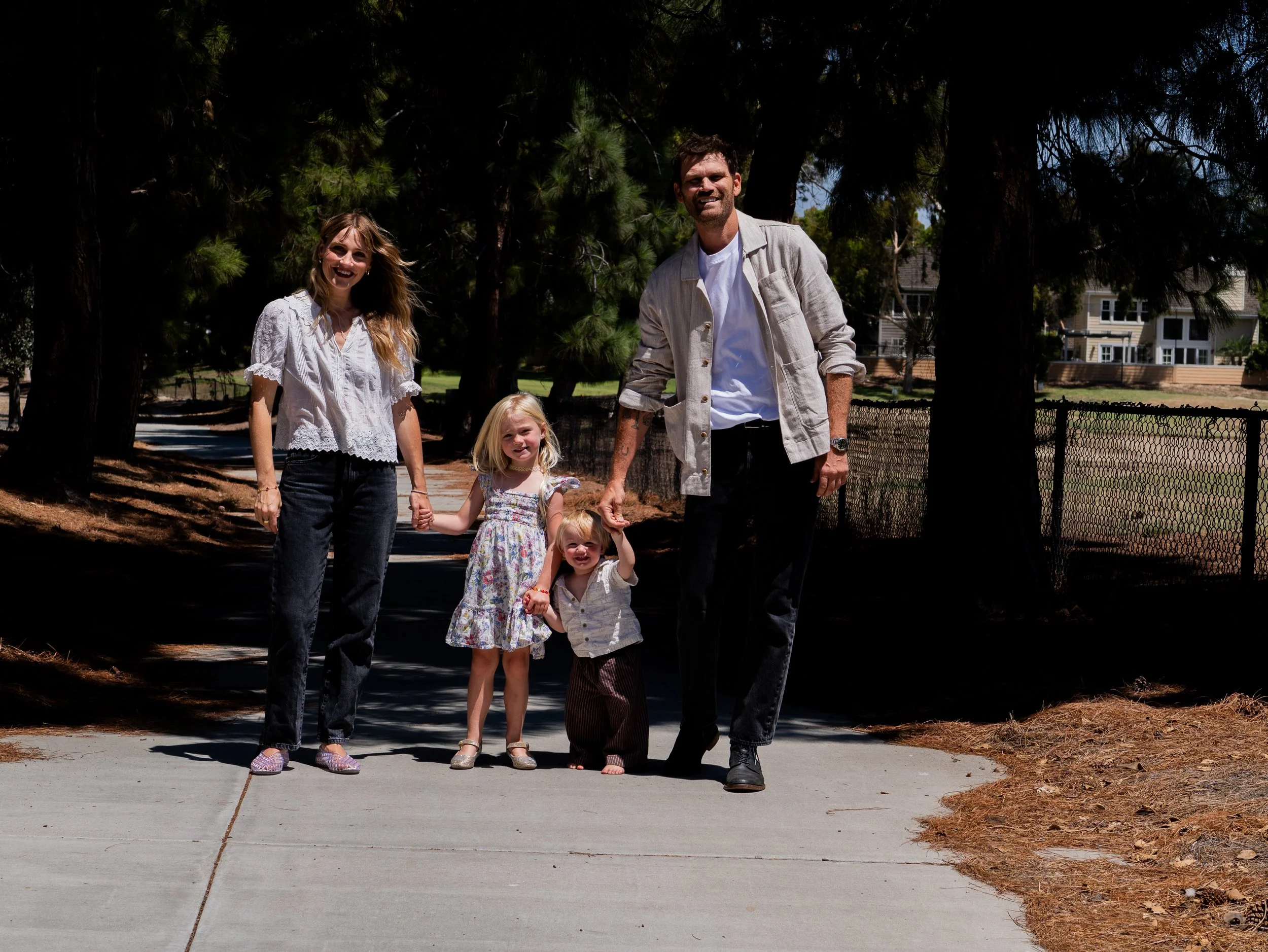 A family of four walking on a sidewalk outdoors during daytime, with trees and houses in the background.