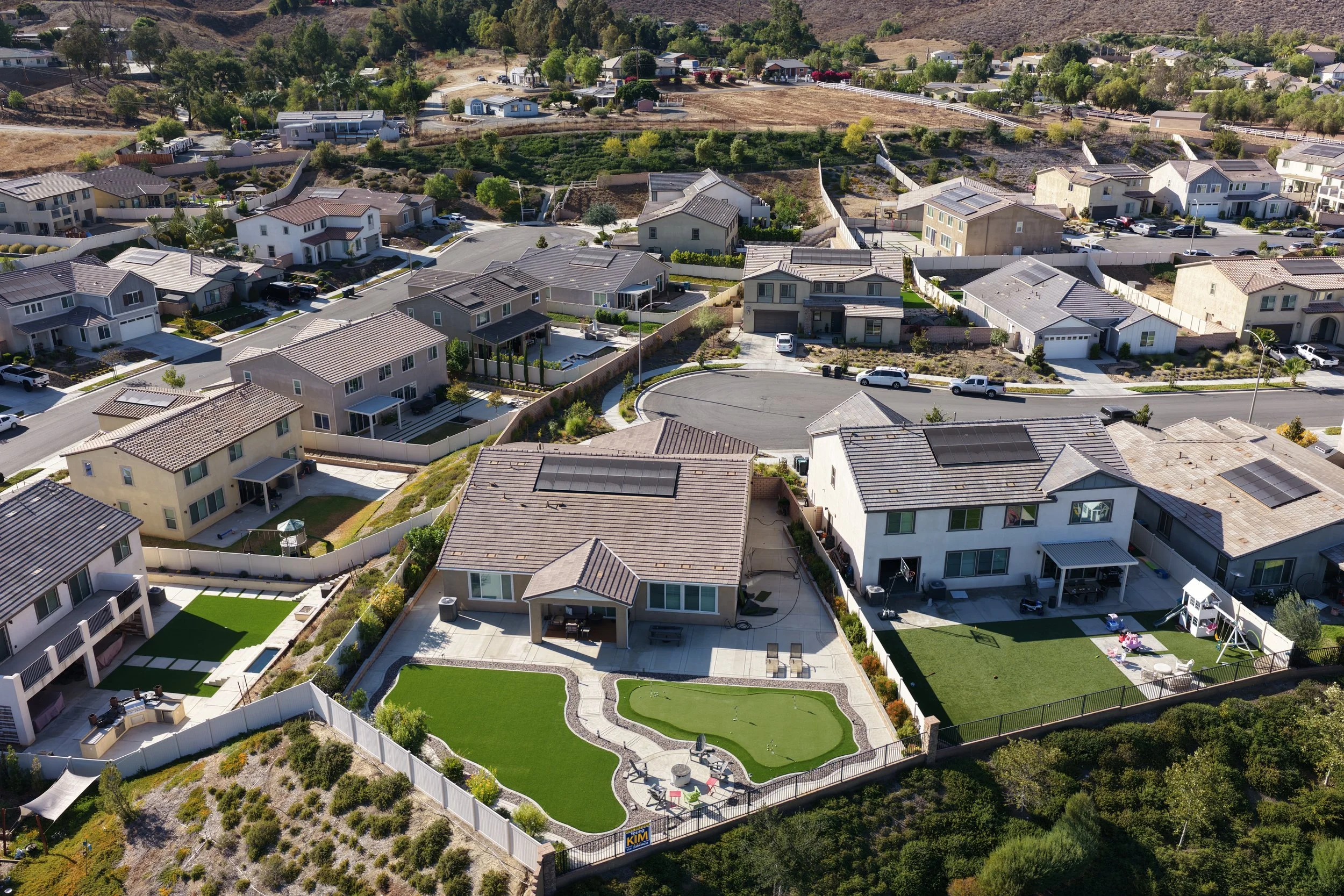Aerial view of a residential neighborhood with single-family homes, fences, yards, and a small public park with a putting green, seating, and playground equipment.