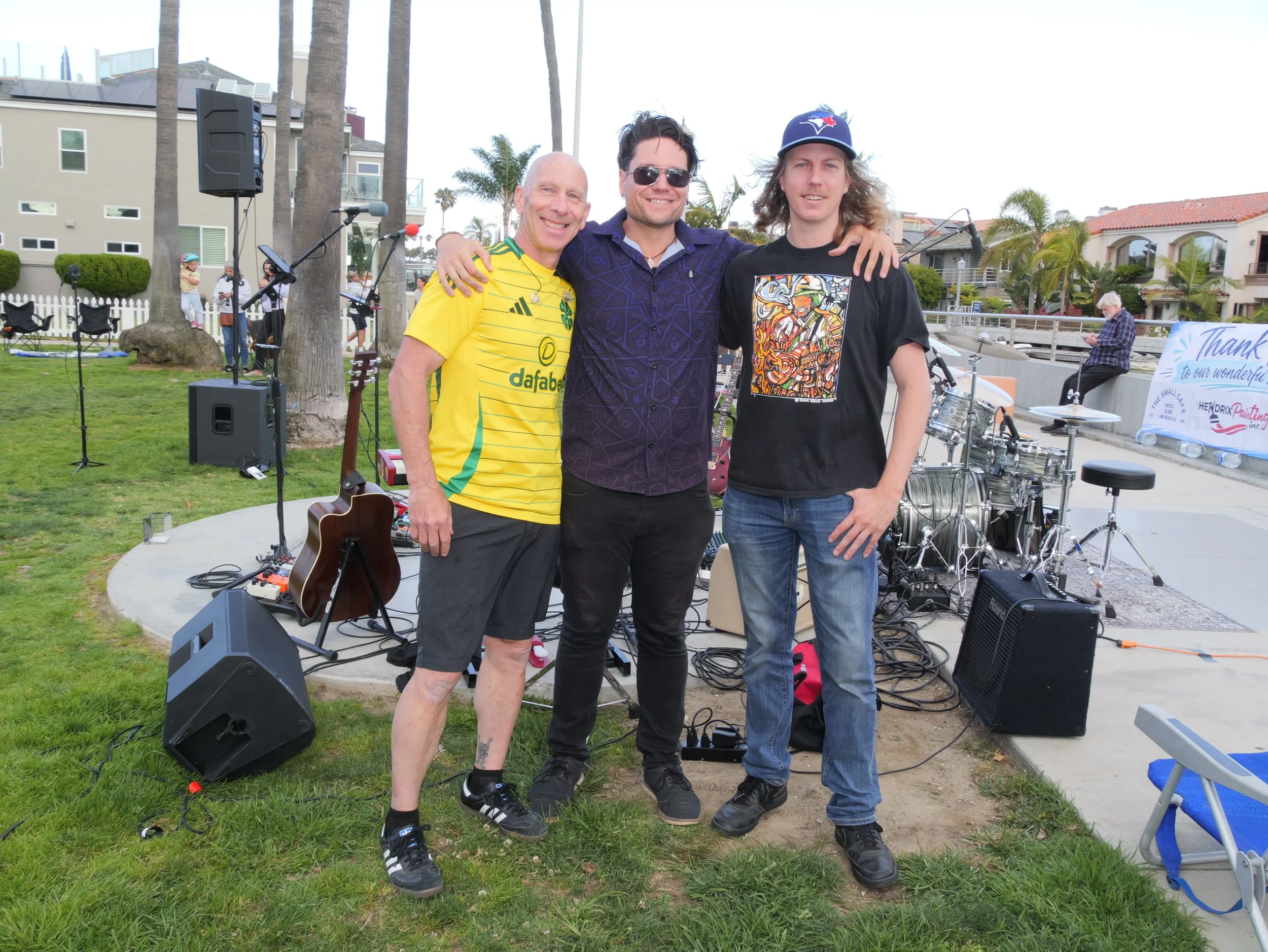 Three men standing closely together outdoors with their arms around each other's shoulders, smiling at the camera. They are at a backyard event with musical equipment set up, palm trees, and a white fence in the background.