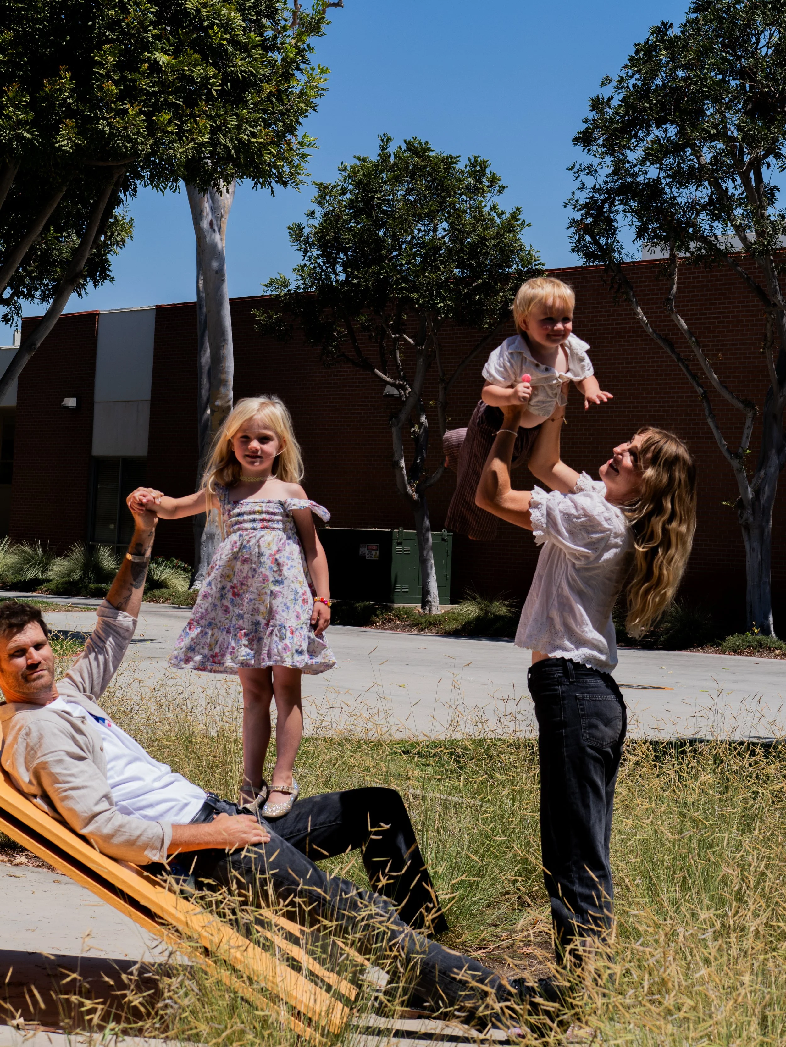 A woman is lifting a smiling toddler girl into the air outdoors near trees and a brick building. A young girl in a floral dress stands nearby, holding hands with a man sitting in a lounge chair, all enjoying a sunny day.