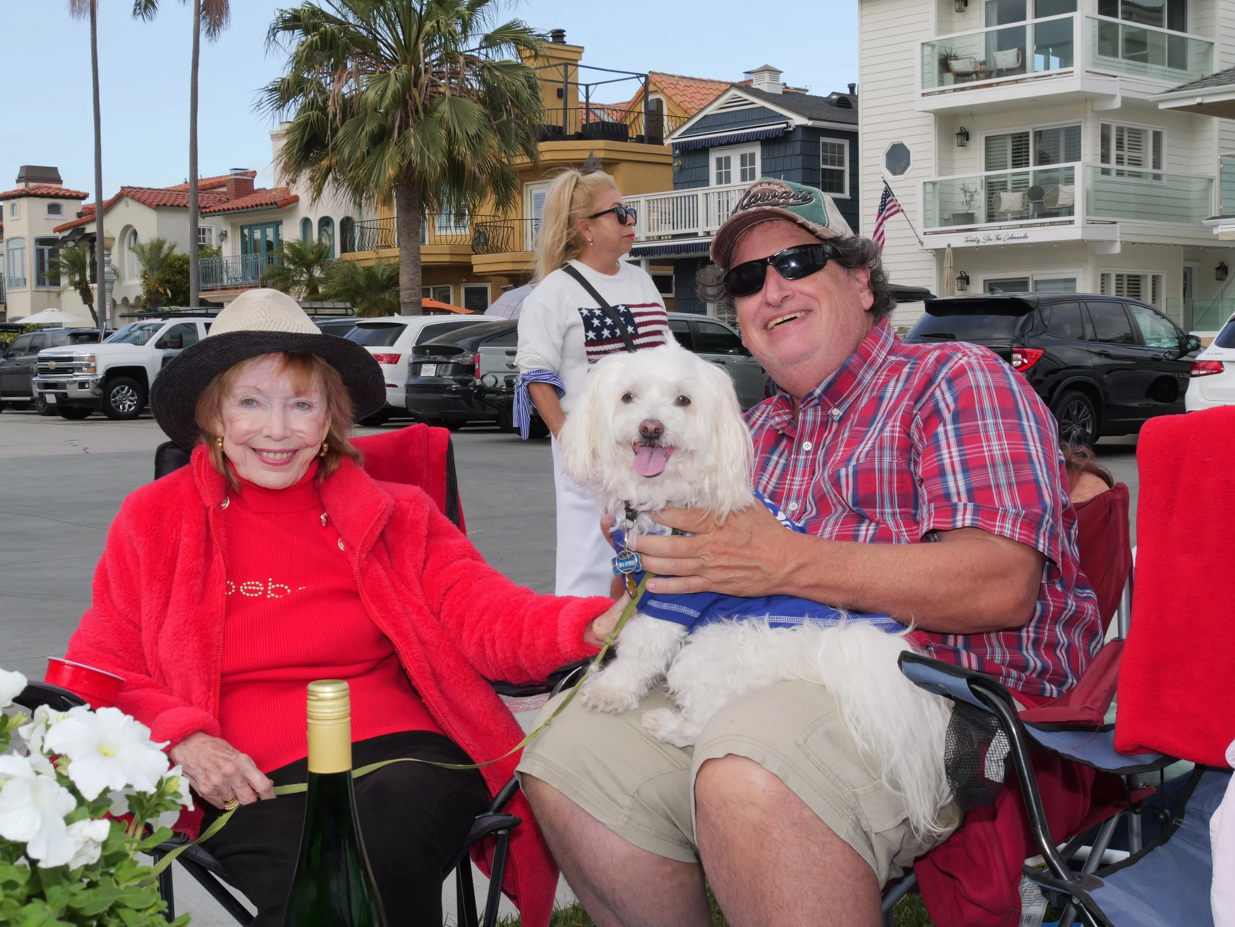 Group of people celebrating outdoors with a dog, in front of colorful houses and palm trees, with a table holding flowers and wine bottles.