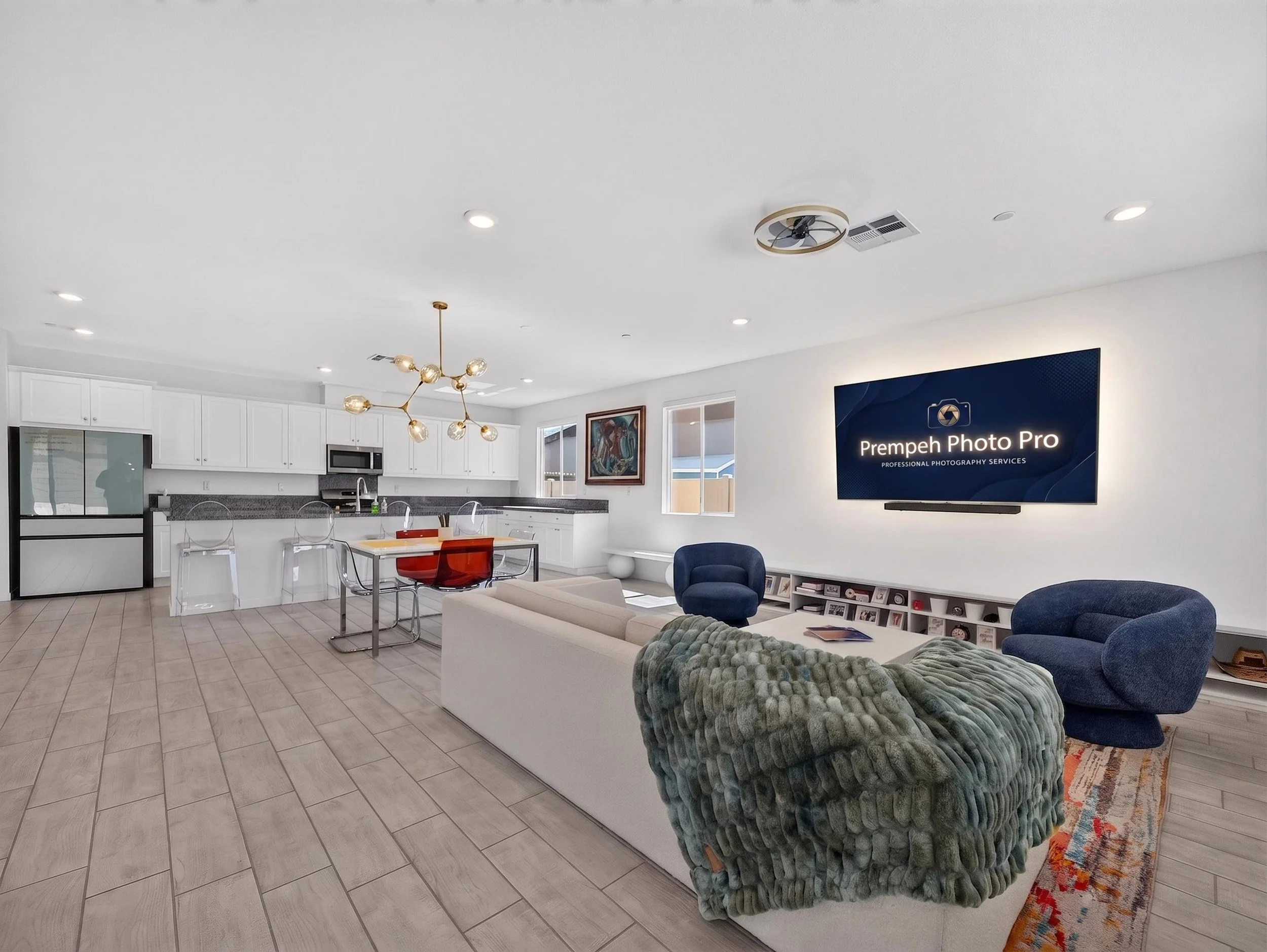 A modern living room with a kitchen in the background, featuring a round dining table with transparent chairs, a velvet green armchair, a blue armchair, a large wall-mounted television with a photo pro logo, and decorative artwork on the walls. The r