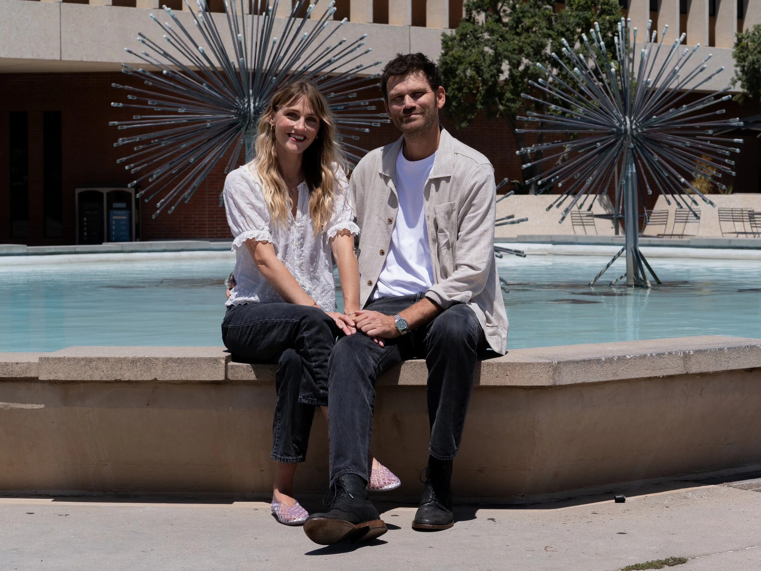 A young woman with long blonde hair and a young man with dark hair and a beard sitting closely together on a low wall in front of a modern building and artistic metal sculptures, smiling at the camera.