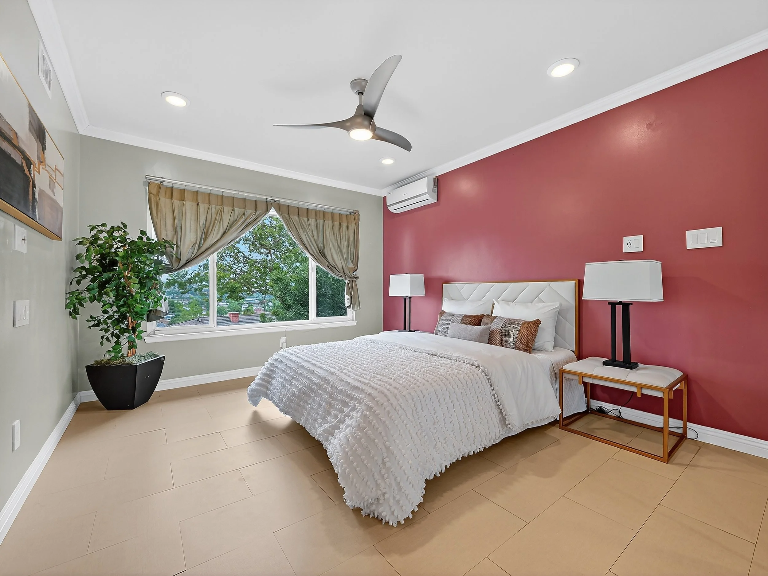 A bedroom with a white bed, pink accent wall, side tables with lamps, a window with beige curtains, a ceiling fan, and a potted plant.