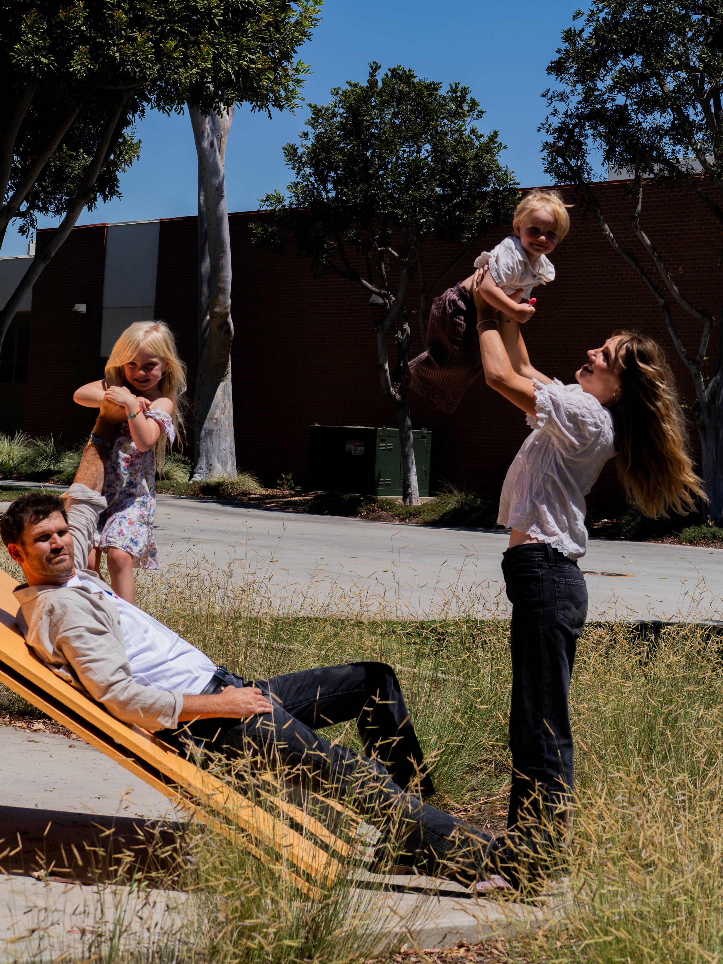A woman with light brown hair is lifting a small child with blonde hair in the air, both are smiling. A girl with blonde hair stands nearby, and a man with dark hair and a beard is lying on a lounge chair, relaxing. They are outdoors with trees, gras