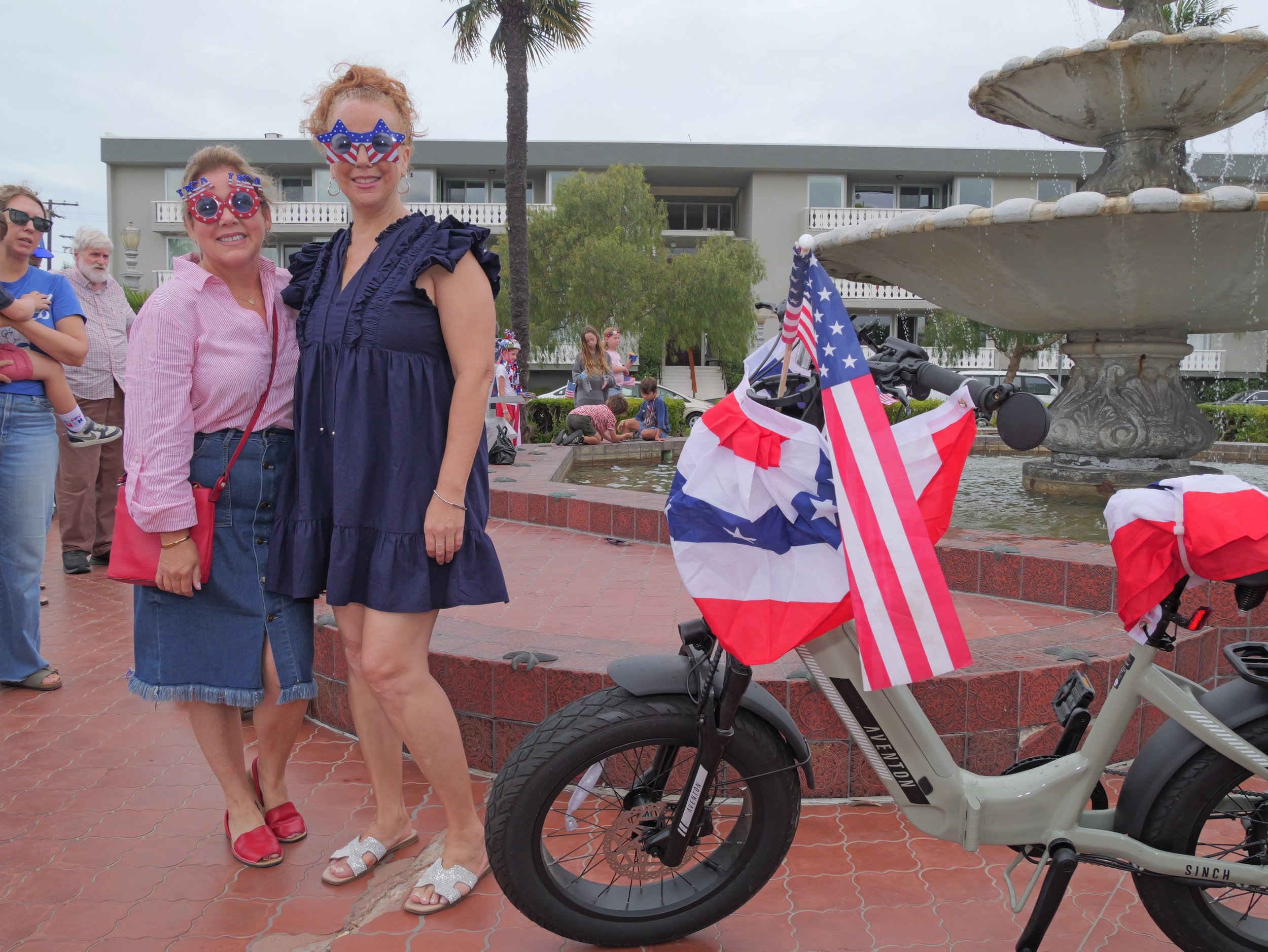 Two women at a patriotic celebration wearing American flag themed sunglasses, standing near a fountain with American flags attached to a bike, and a group of people in the background.