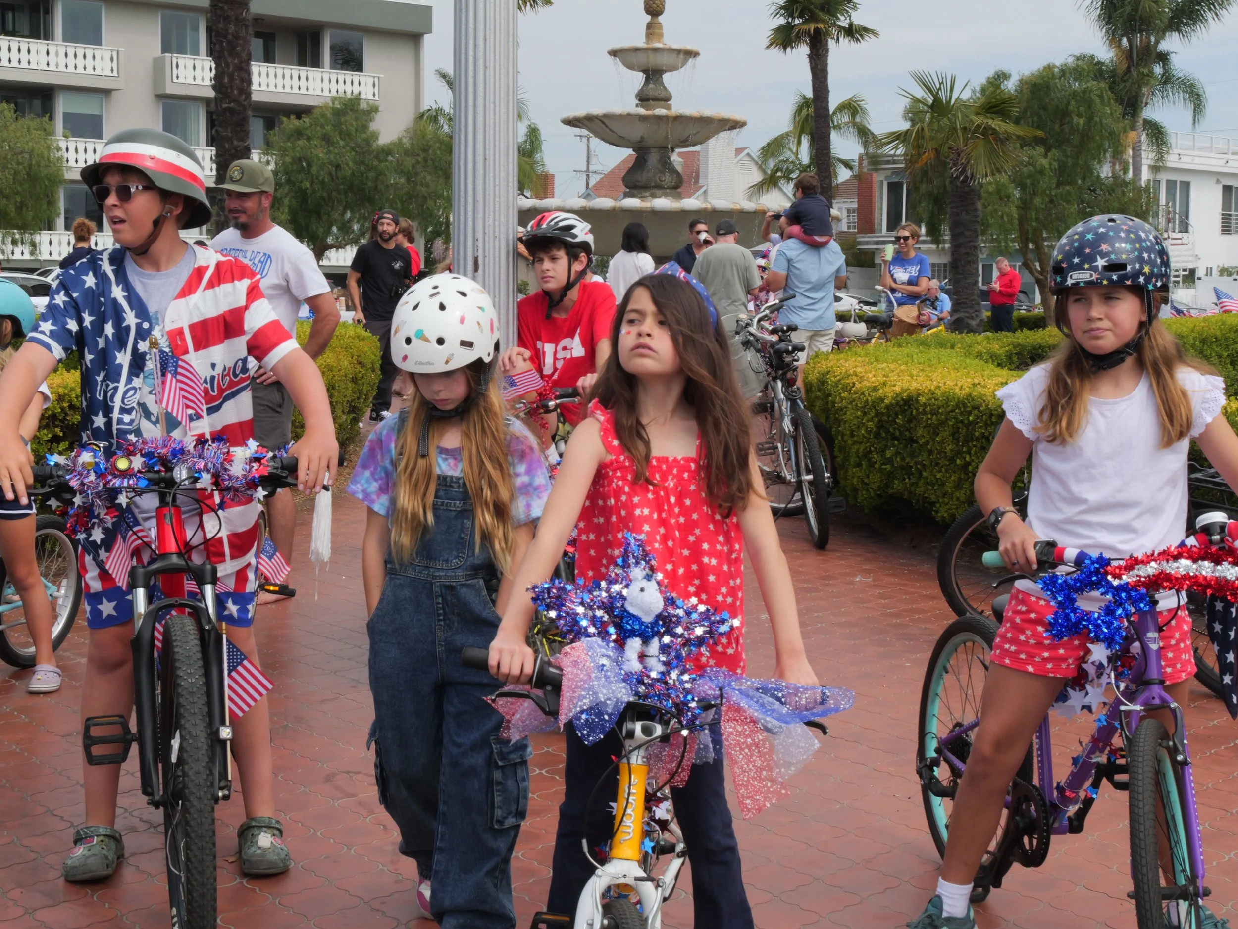 Children participating in a patriotic parade, many wearing red, white, and blue clothing and helmets, riding bikes decorated with American flags and stars, in front of a fountain and palm trees.