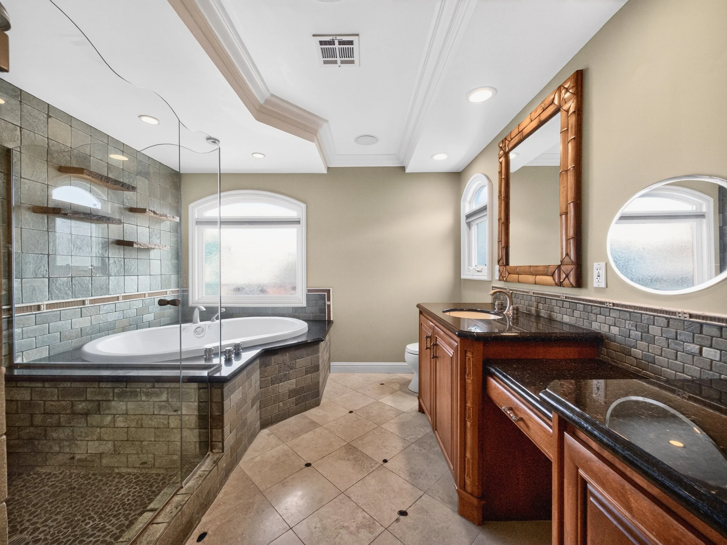 A bathroom with a glass-enclosed shower, a bathtub, a wooden vanity with a black countertop, and a mirror with a bamboo frame. The room has beige walls, white crown molding, and tiled floors.