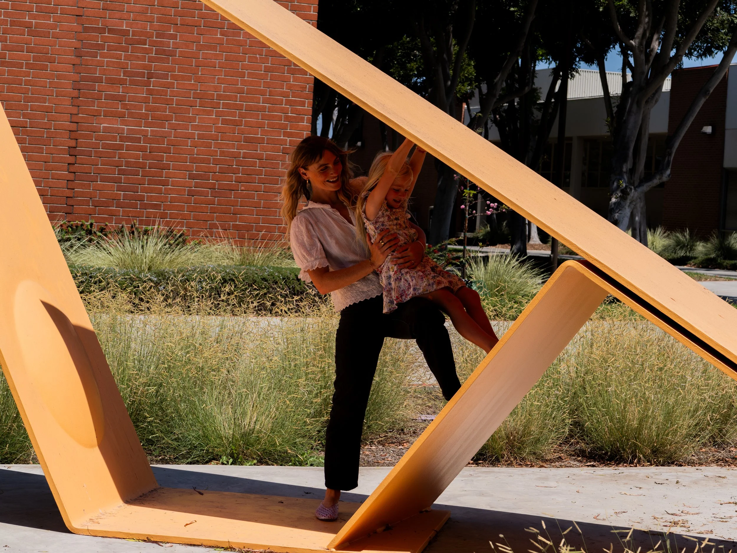 A woman holding a young girl on a large, yellow, abstract outdoor sculpture with brick and trees in the background.