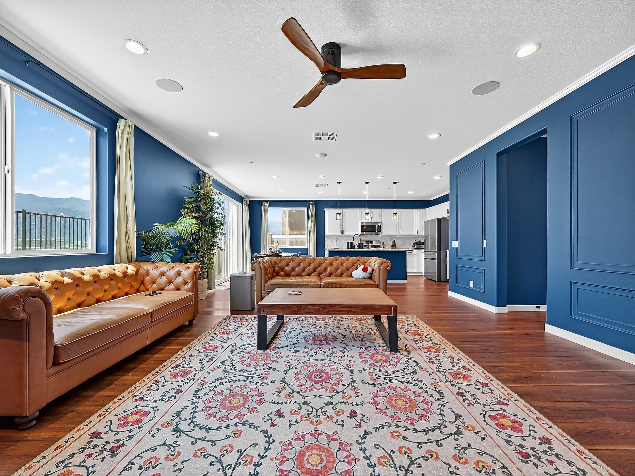 Living room with blue walls, tan leather tufted sofas, a patterned rug, large windows with curtains, and a view of mountains outside.