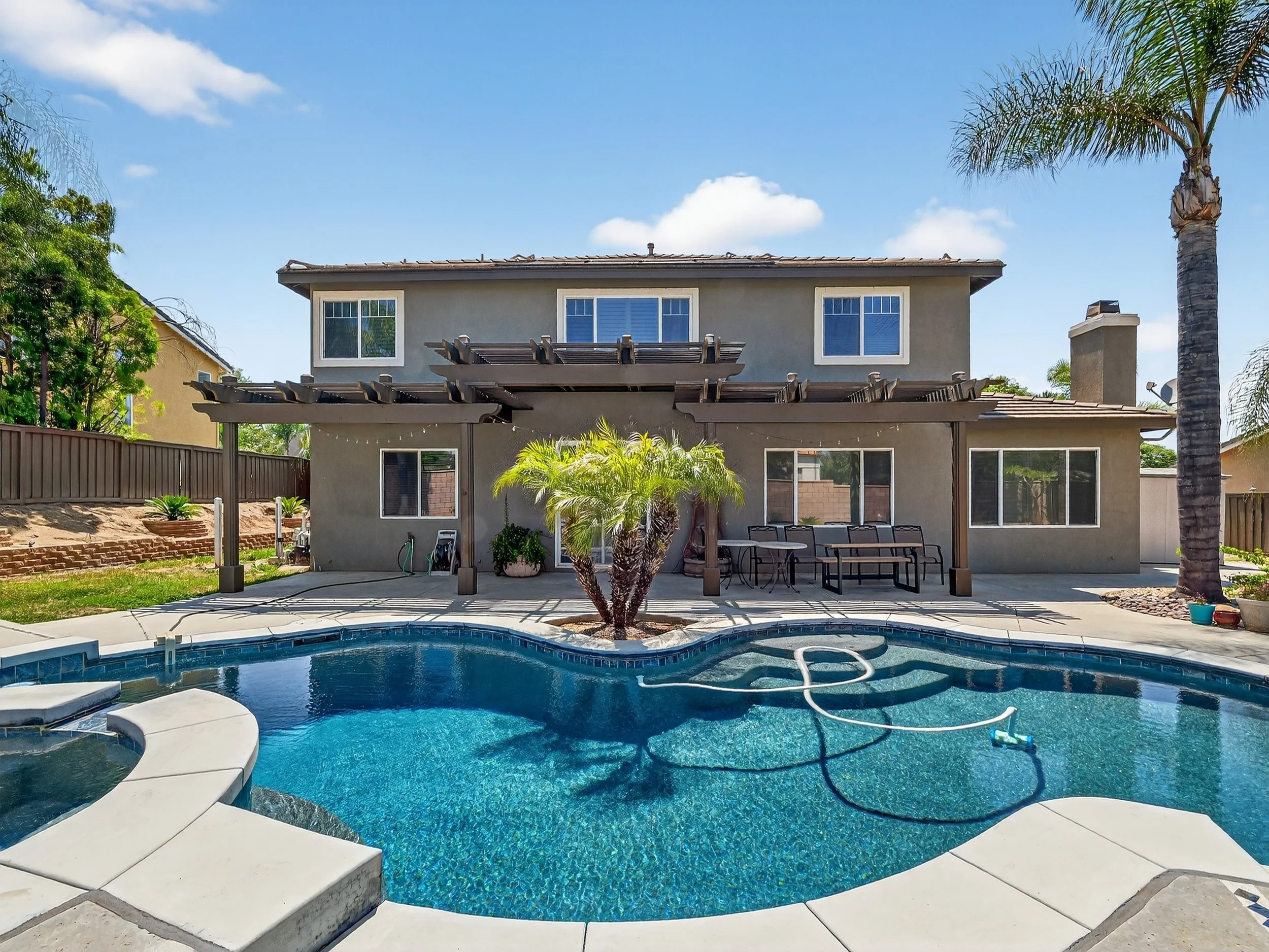 Backyard with a swimming pool, patio chairs, a pergola, and a two-story house with multiple windows and a chimney. There are palm trees and other greenery around.