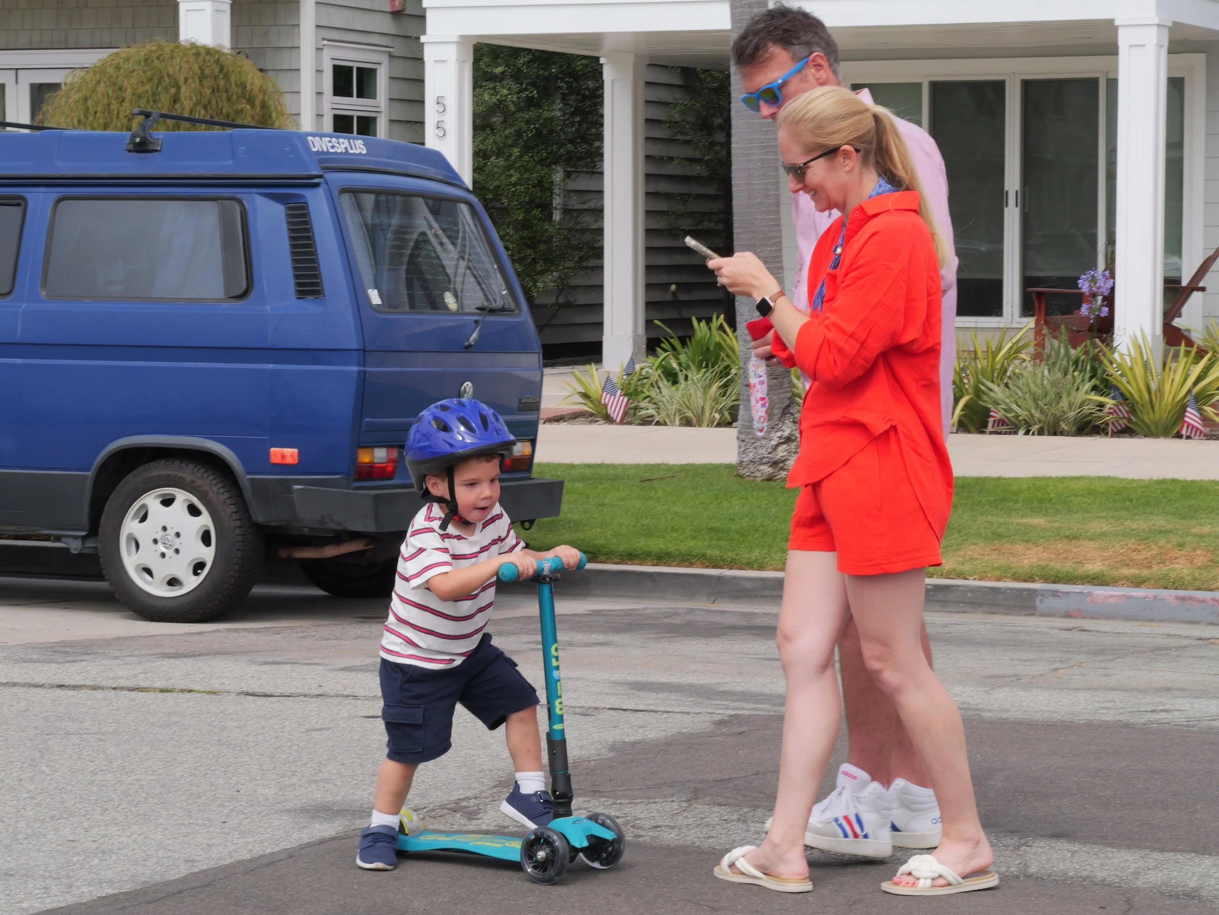 A young boy with a helmet riding a scooter on the street with a woman walking beside him. The boy is wearing a striped shirt and shorts, and the woman is dressed in orange shorts and a blue shirt, wearing glasses. They are in front of a house with fl
