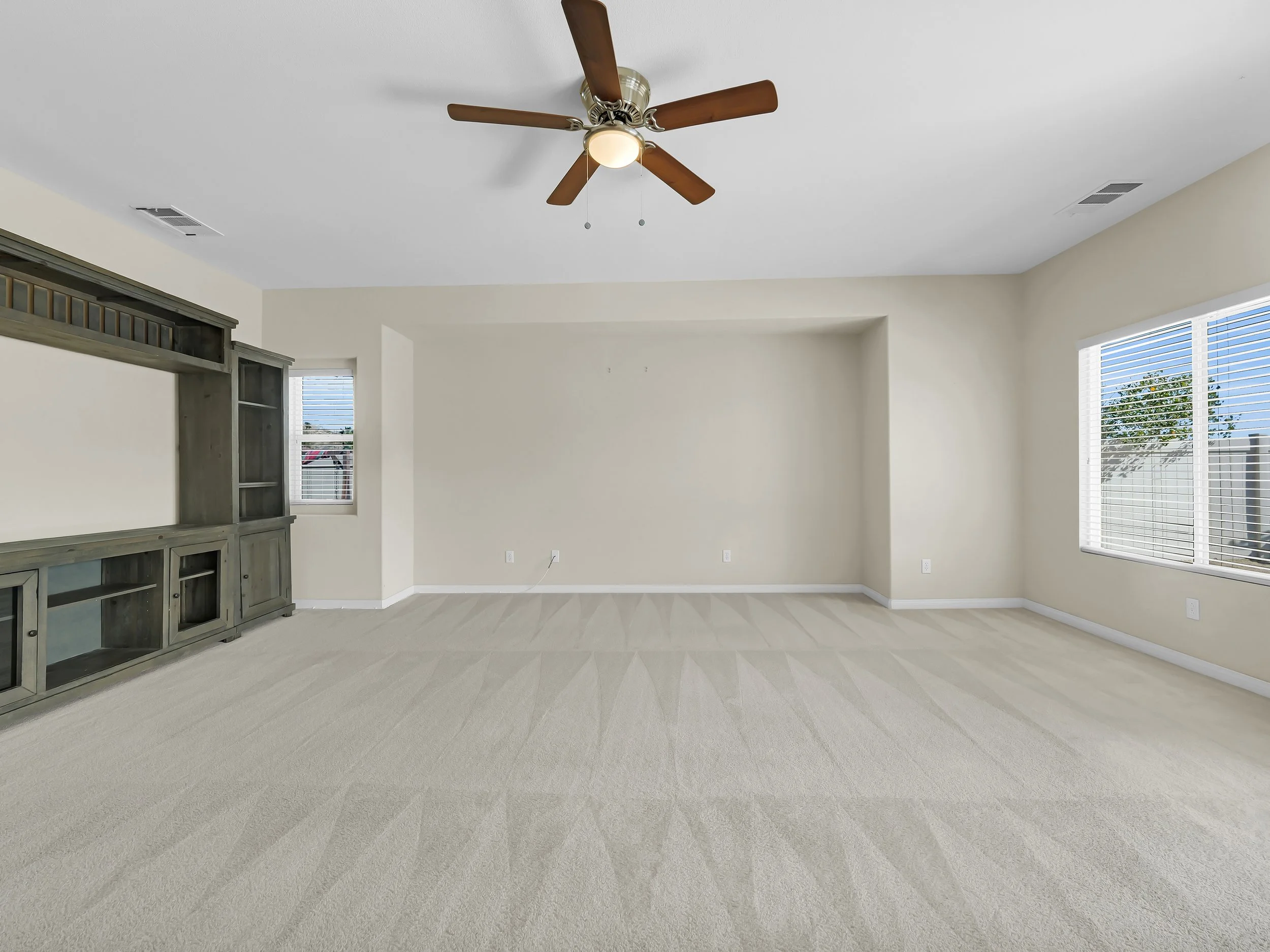Empty living room with beige carpet, white walls, large windows with blinds, ceiling fan, and a gray wooden bookshelf.