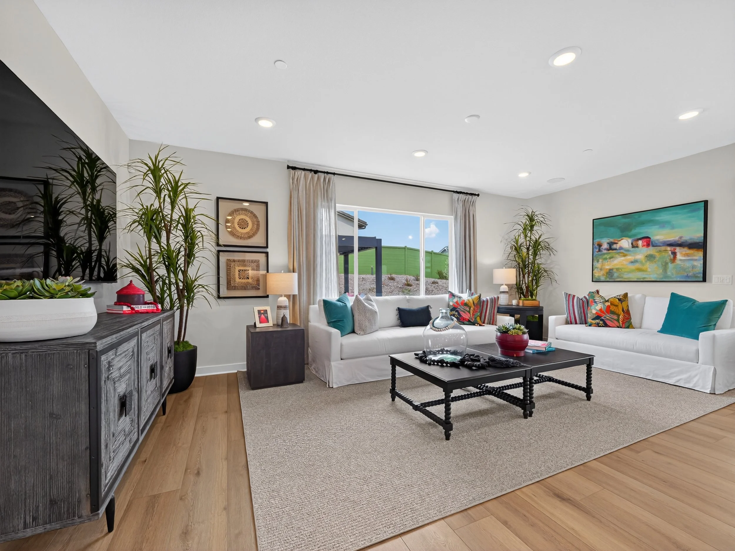 Living room with white sofas, colorful pillows, coffee table, wall-mounted artwork, large window with curtains, indoor plants, and a view of the backyard.
