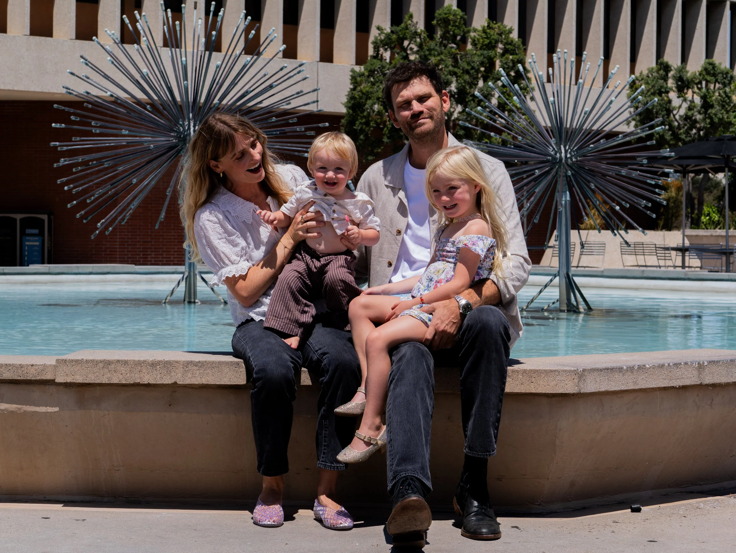 A family of four sitting on a concrete ledge in front of a fountain with artistic metal sculptures, smiling and enjoying a sunny day outside in an urban setting.