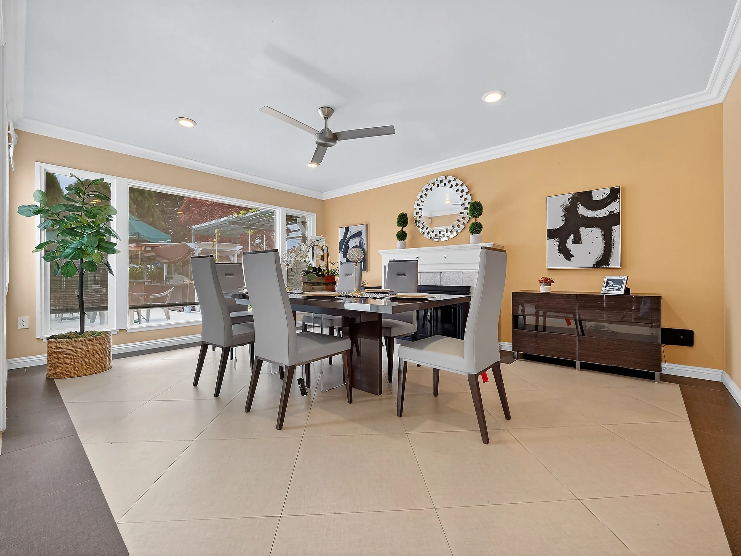 Bright dining room with white ceiling, recessed lighting, ceiling fan, large windows, beige walls, modern abstract art, potted plant, and a dark wood dining table with white chairs.