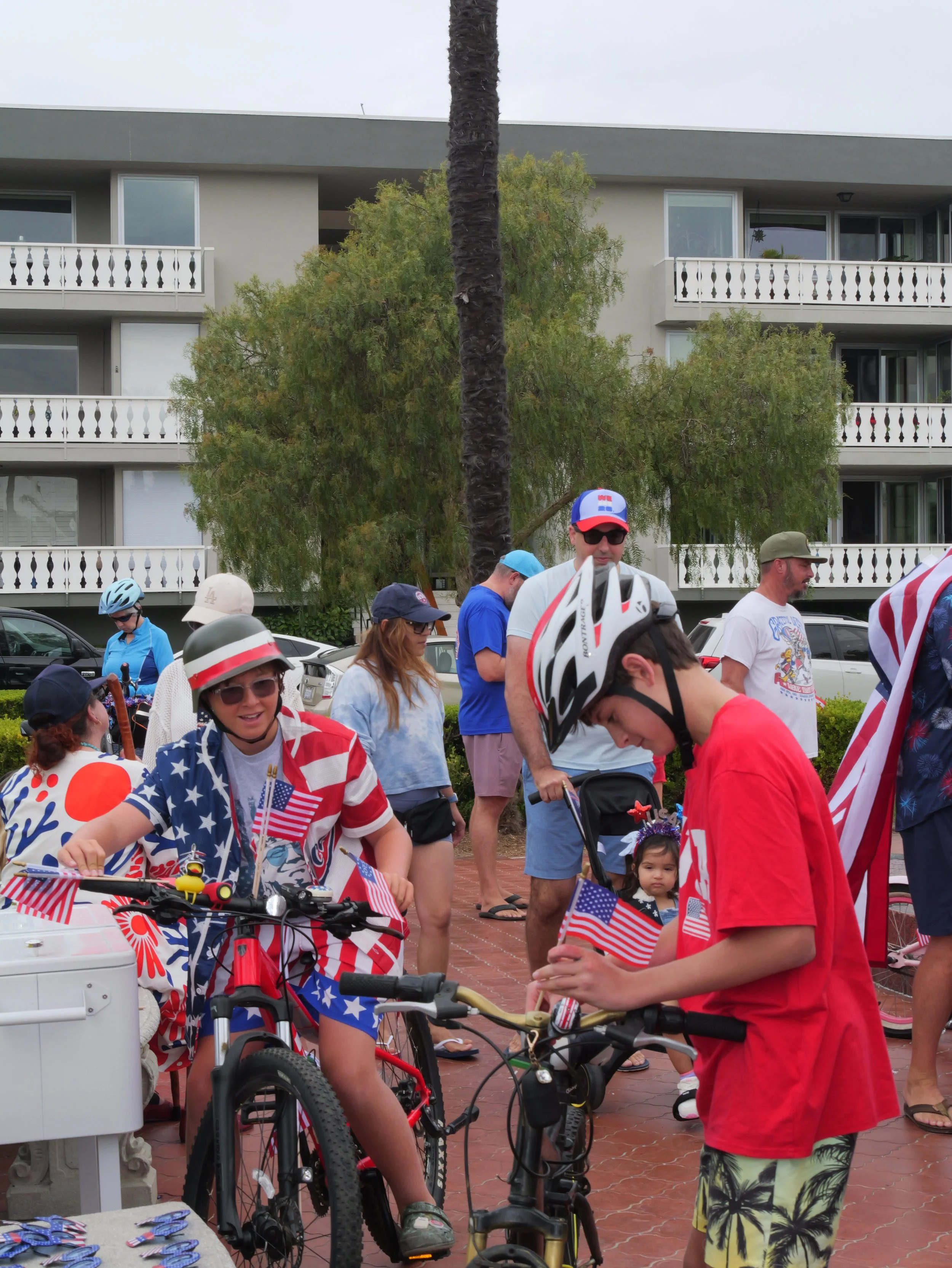 People gathered outdoors during a celebration, many with American flags and helmets, some standing near bicycles, with a building and trees in the background.