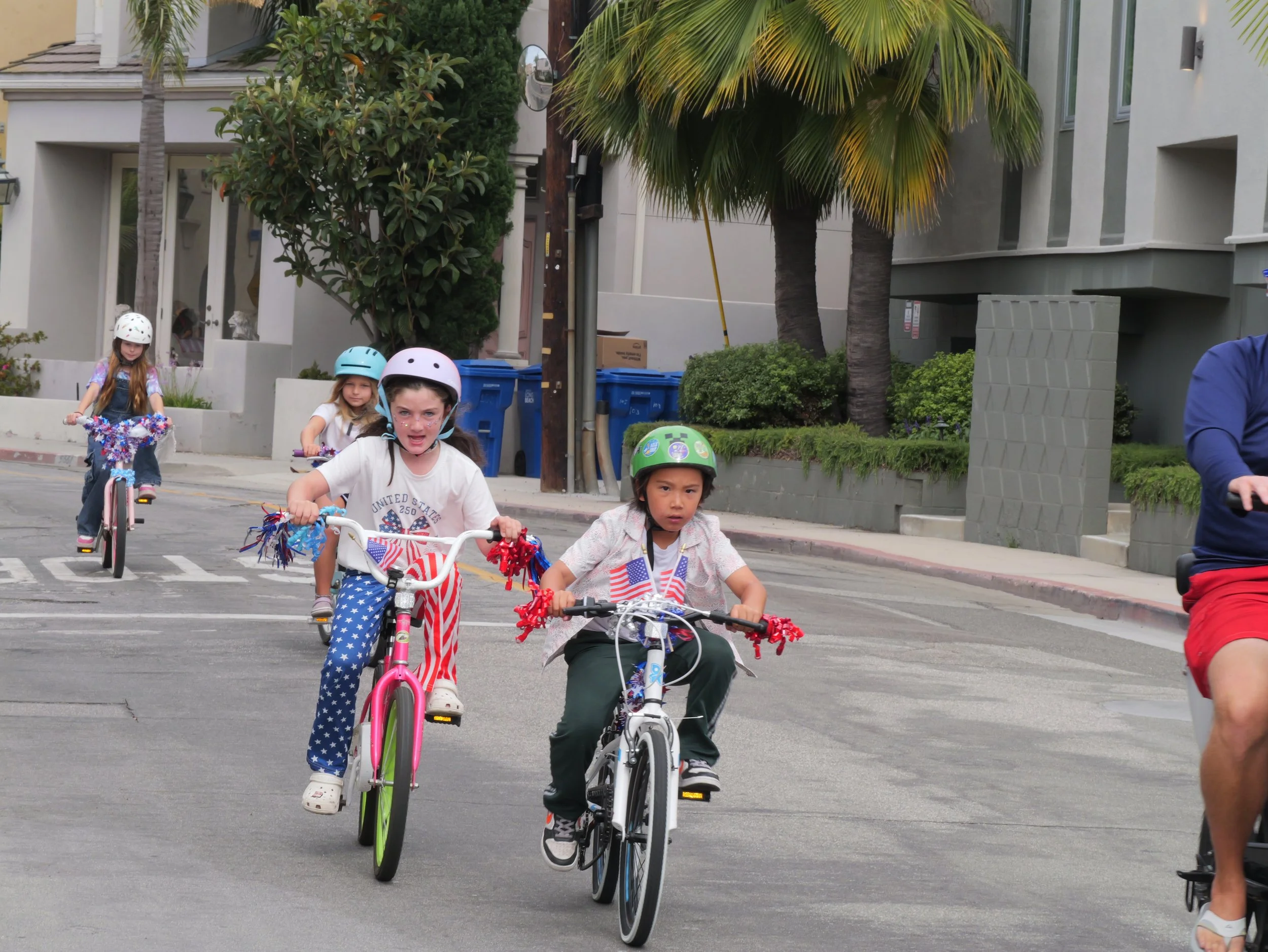 Children riding bicycles with helmets on a city street during a parade or celebration, with palm trees and modern buildings in the background.