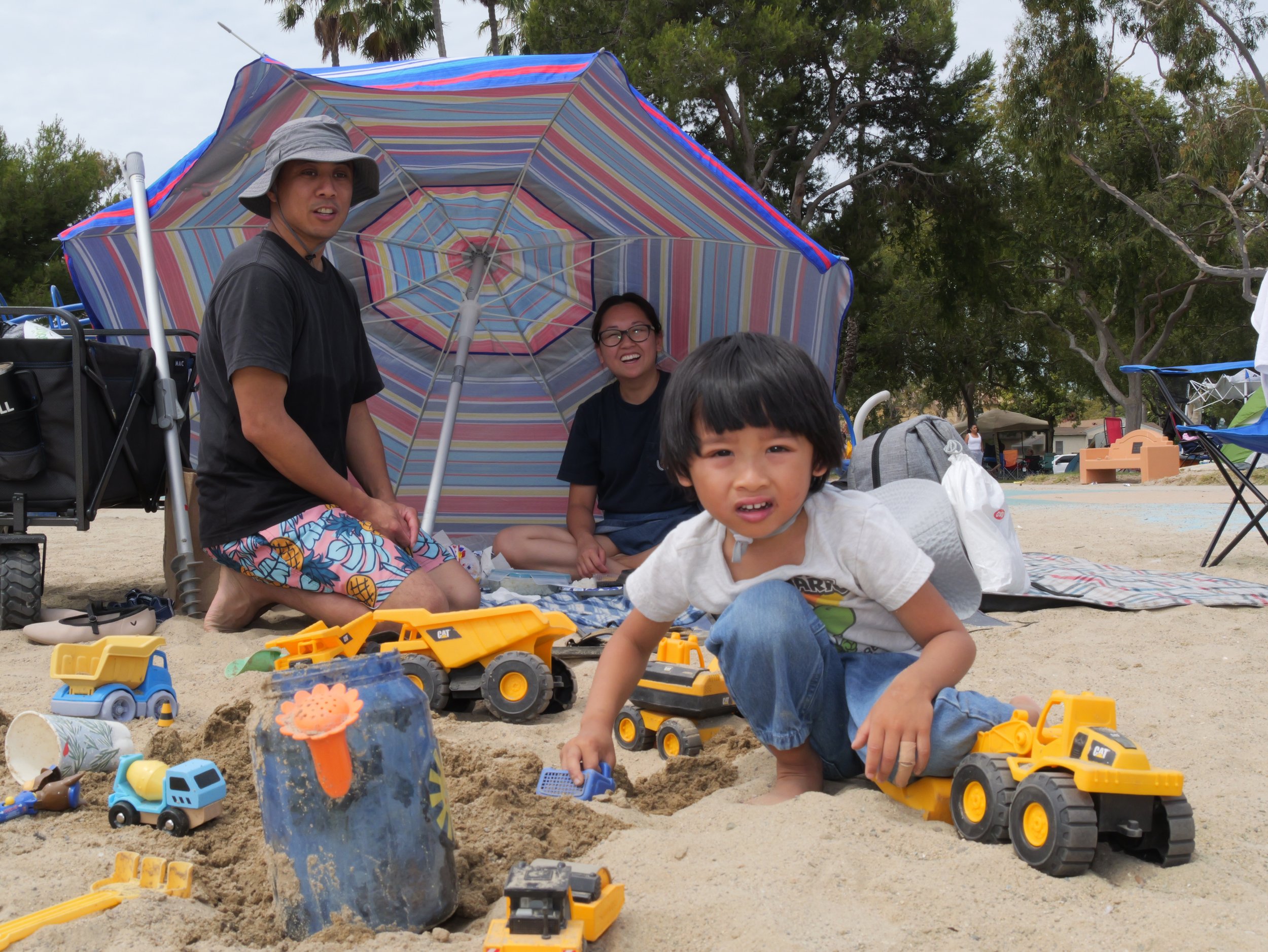 People at the beach playing in the sand with toy trucks and containers, under an umbrella with trees in the background.