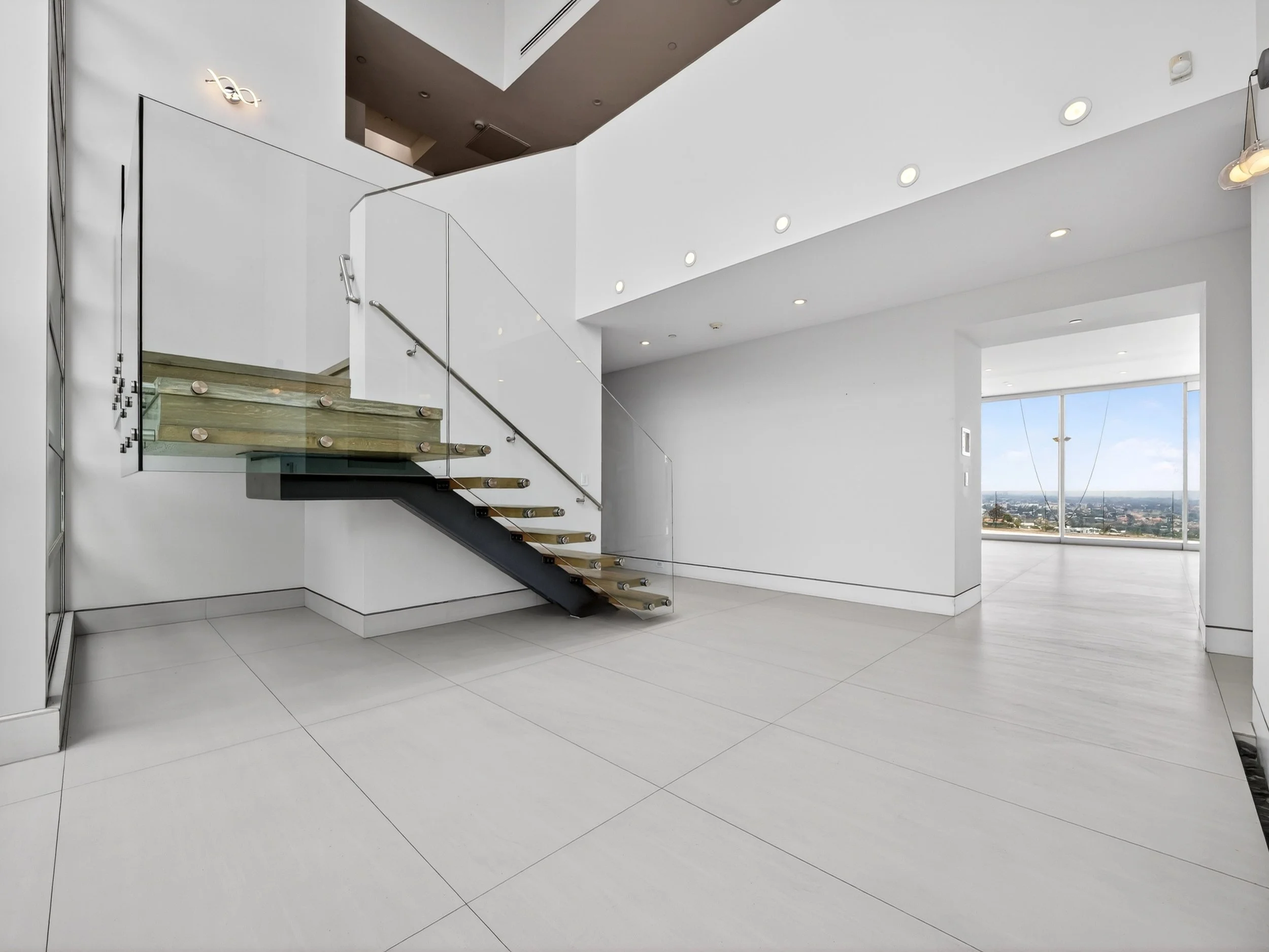 Modern minimalist interior with white walls, a staircase with glass railing, and large windows showing a cityscape view.