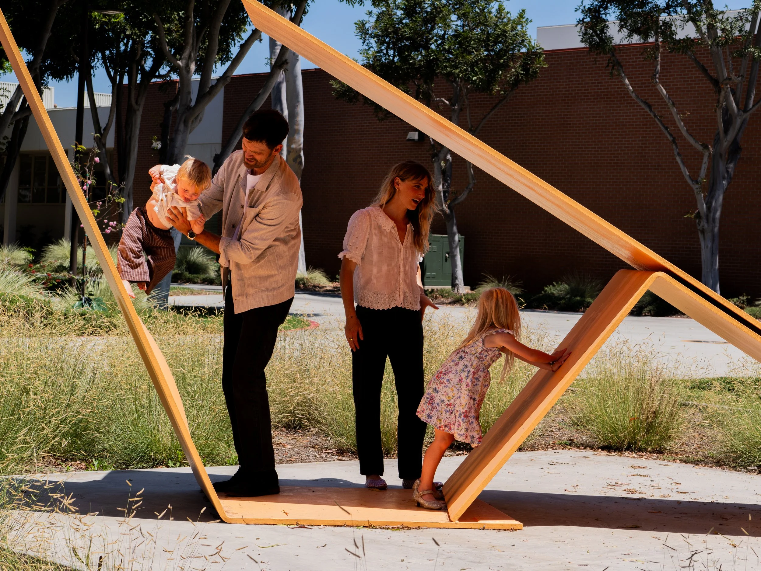 A group of children and a man are playing under a large wooden frame outdoors on sunny day. Children are trying to crawl through the frame while an adult helps a young child over it. A woman observes nearby, smiling. Trees and a brick building are in