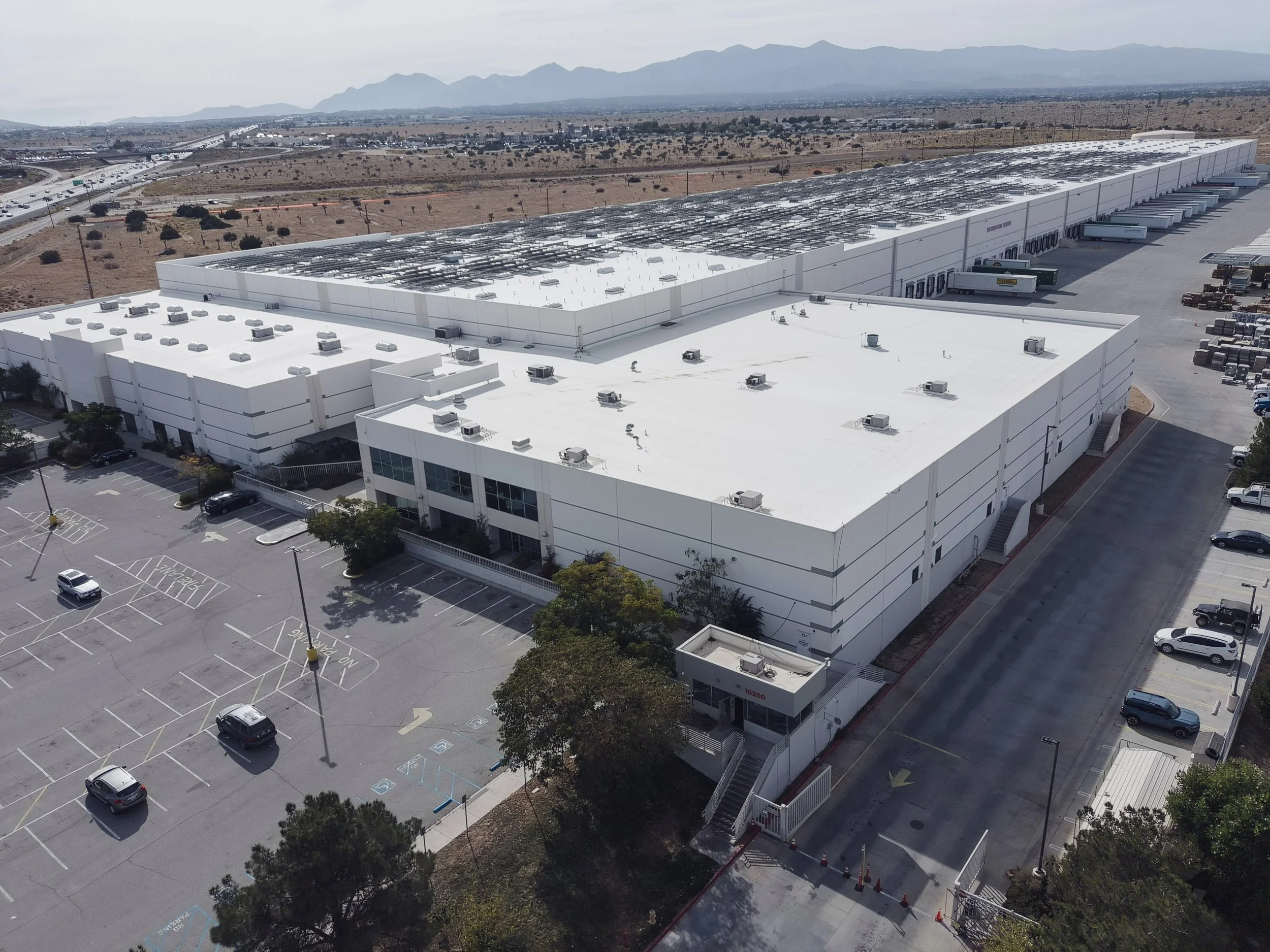 An aerial view of a large white commercial building with solar panels on the roof, a parking lot with some cars, and a distant highway in a desert landscape with mountains in the background.