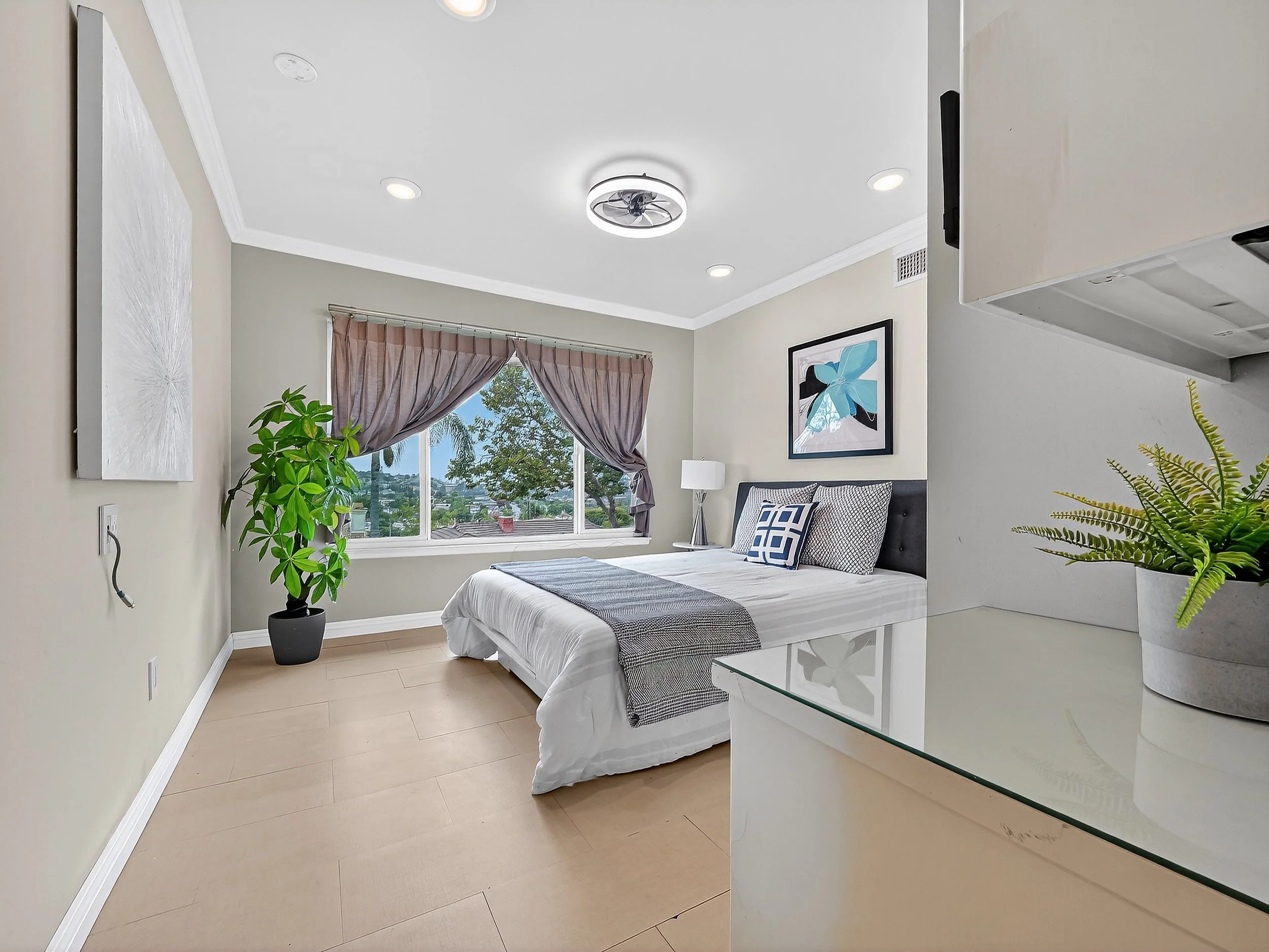 A bedroom with beige walls and light wood flooring, featuring a bed with a black headboard, white bedding, and black-and-white pillows. There are two nightstands with lamps beside the bed. Decorative artwork hangs above the bed, and large windows with brown curtains reveal trees outside. Several green plants are placed around the room, and the ceiling has a modern circular fan with lighting.