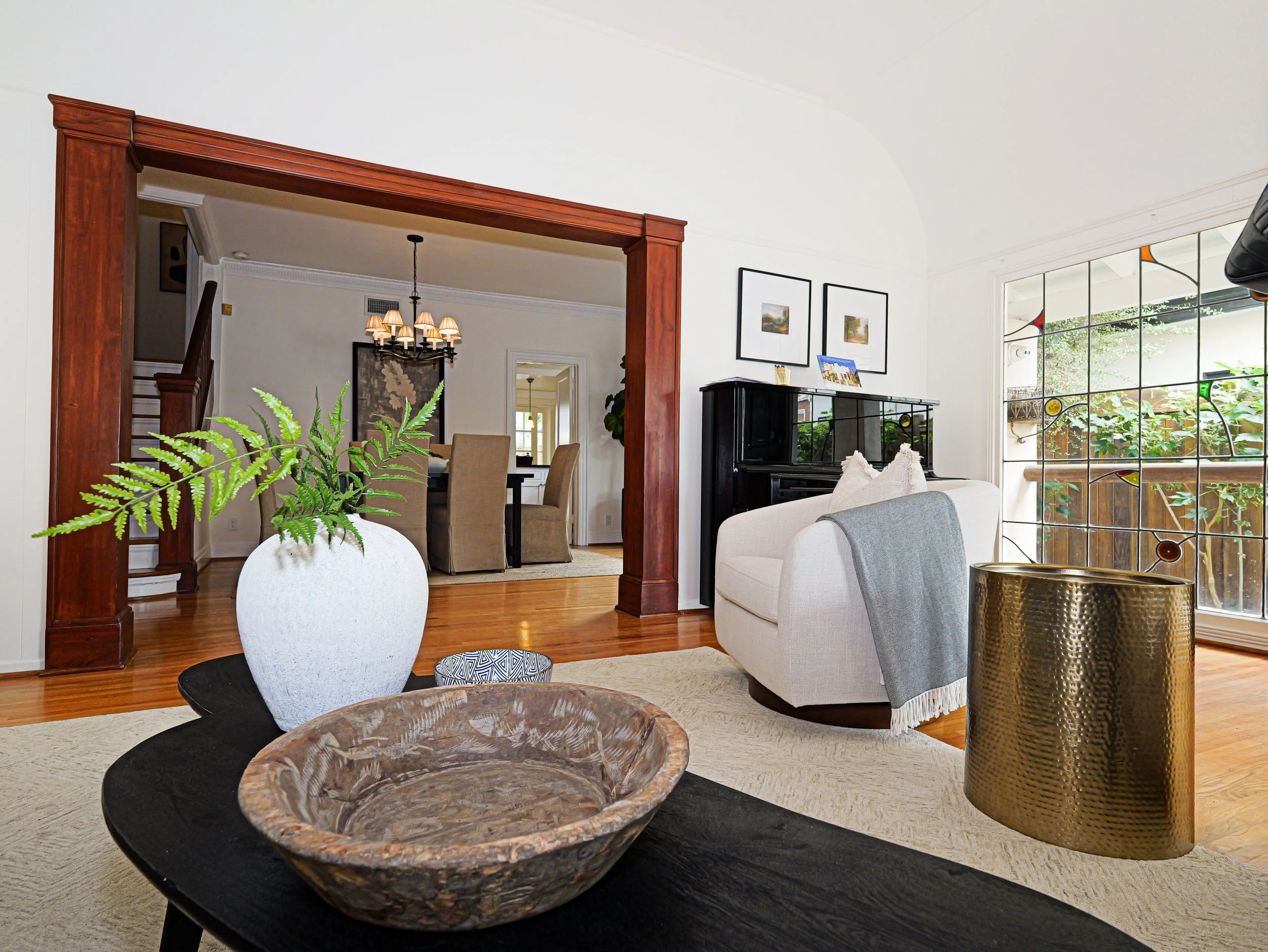 Living room with white walls, a large window with stained glass accents, a beige sofa with white pillows, a black piano, three framed pictures on the wall, and a green potted plant in a white pot. There are wooden floors and a chandelier in a dining 