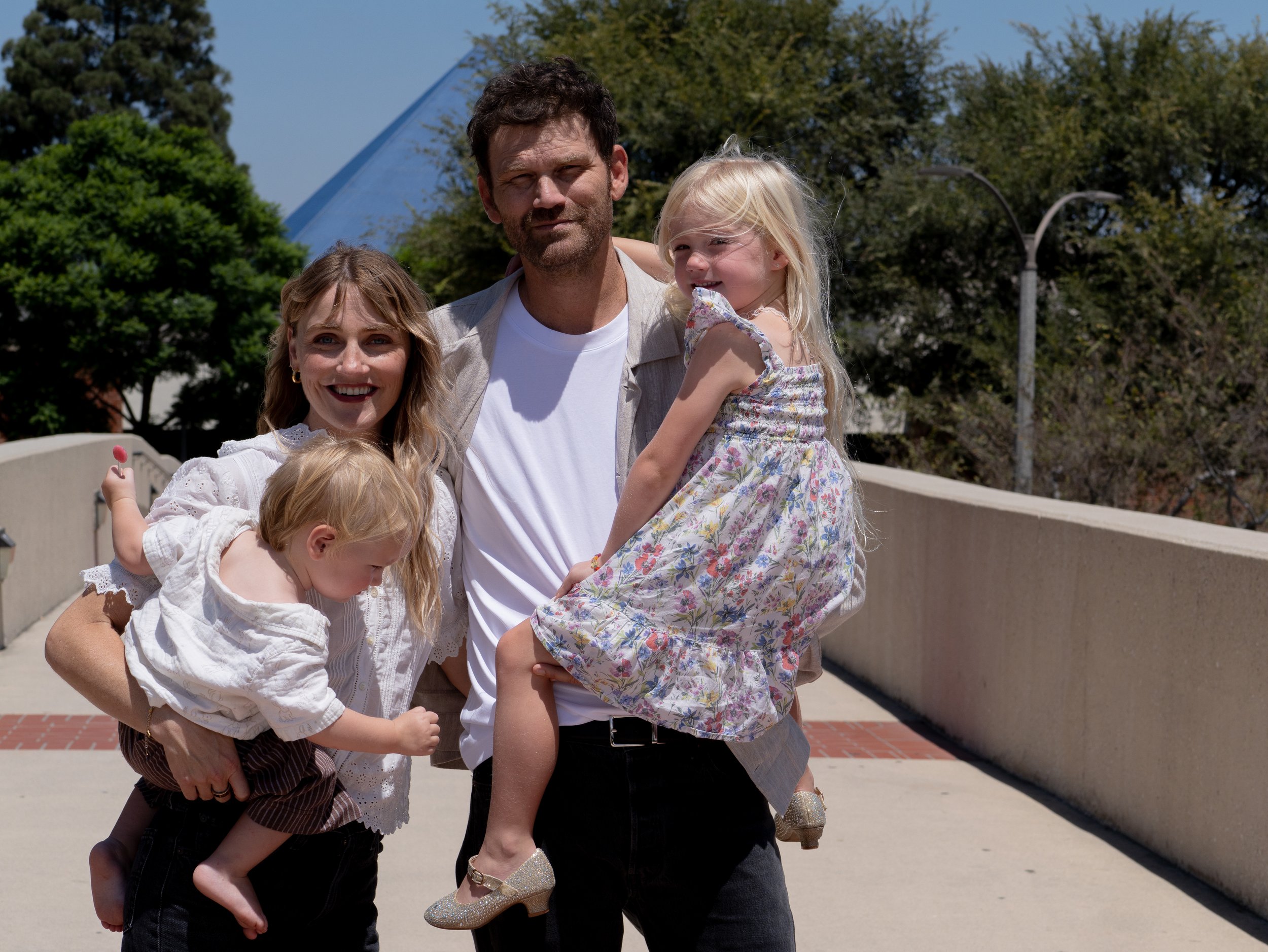A group of five people, a man, woman, and three young girls, posing outdoors on a sunny day with trees and a building in the background. The man is carrying two of the girls, one on each side, and they are all smiling or looking at the camera.