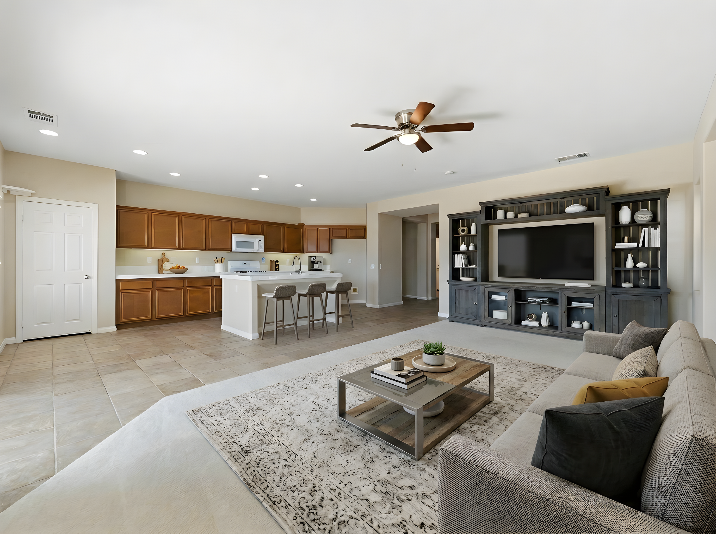 Open-concept living room with beige sofa, coffee table, and built-in dark gray shelving with TV, adjacent to kitchen with wooden cabinets, white countertops, and bar stools. Ceiling fan and recessed lighting.
