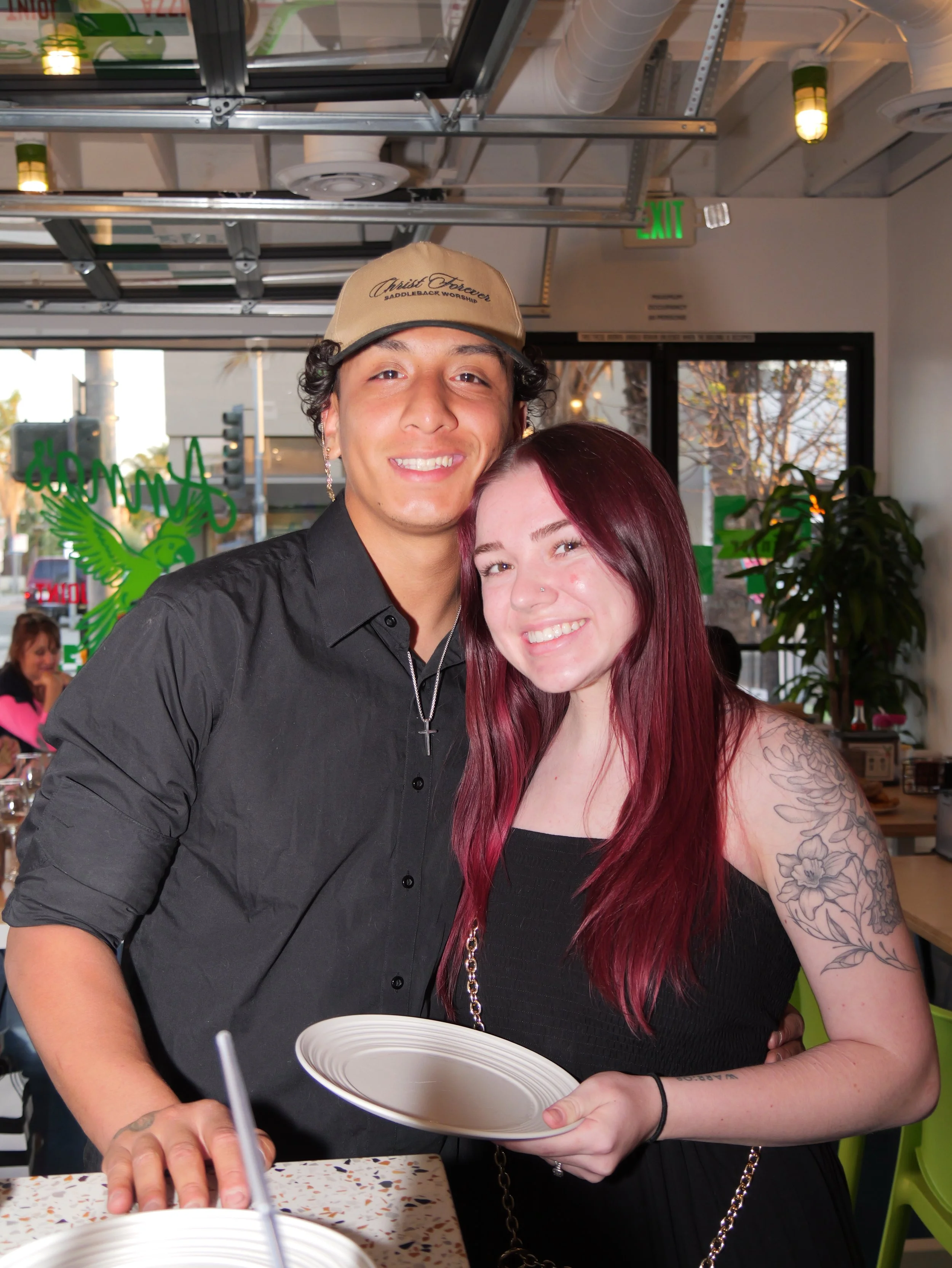 A young man and woman are smiling and looking at the camera inside a restaurant or cafe. The man is wearing a beige cap and a black shirt with a cross necklace. The woman has long, red hair and a floral tattoo on her right arm, and is wearing a black