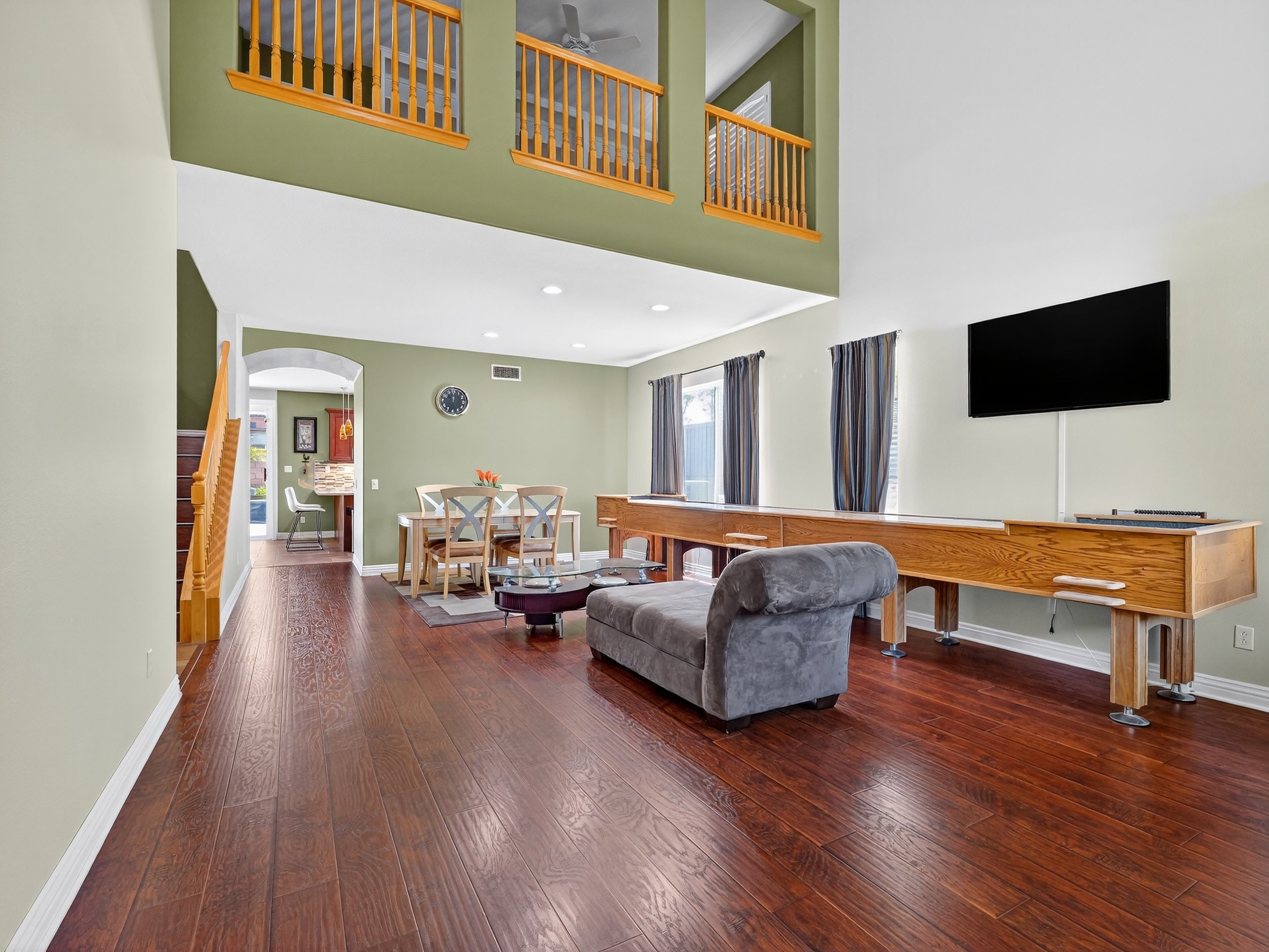 Open-concept living and dining area with green walls, hardwood flooring, a ceiling fan, a wall-mounted TV, a dining table with chairs, and a kitchen in the background.