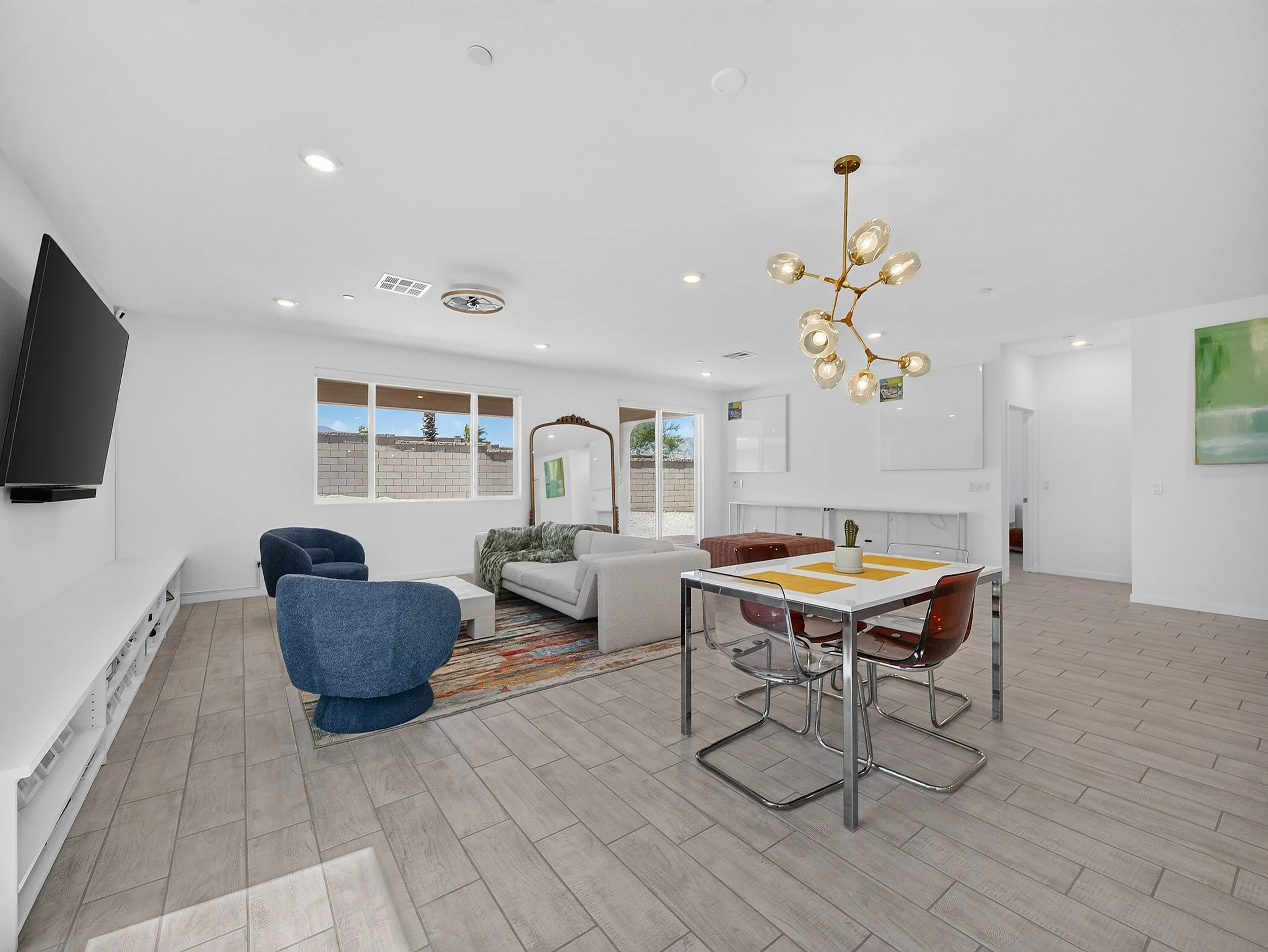 Modern living room with white walls, large windows, a wall-mounted flat-screen TV, blue and white seating, a patterned area rug, a gold chandelier, a dining table with chairs, and artwork on the wall.
