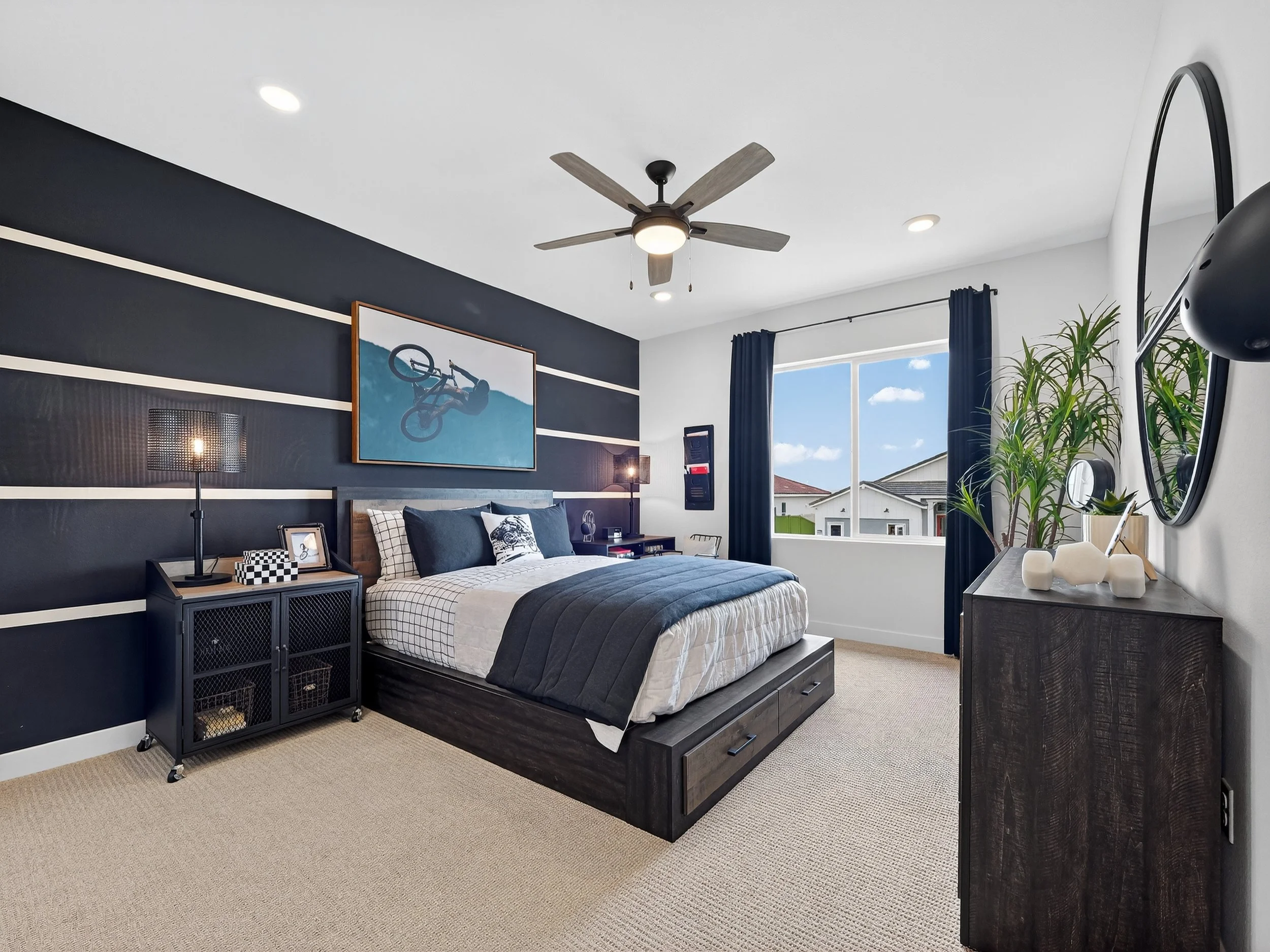 A modern bedroom with a large bed, dark furniture, a ceiling fan, large window with curtains, and wall art. There are plants and decorative objects on the furniture.