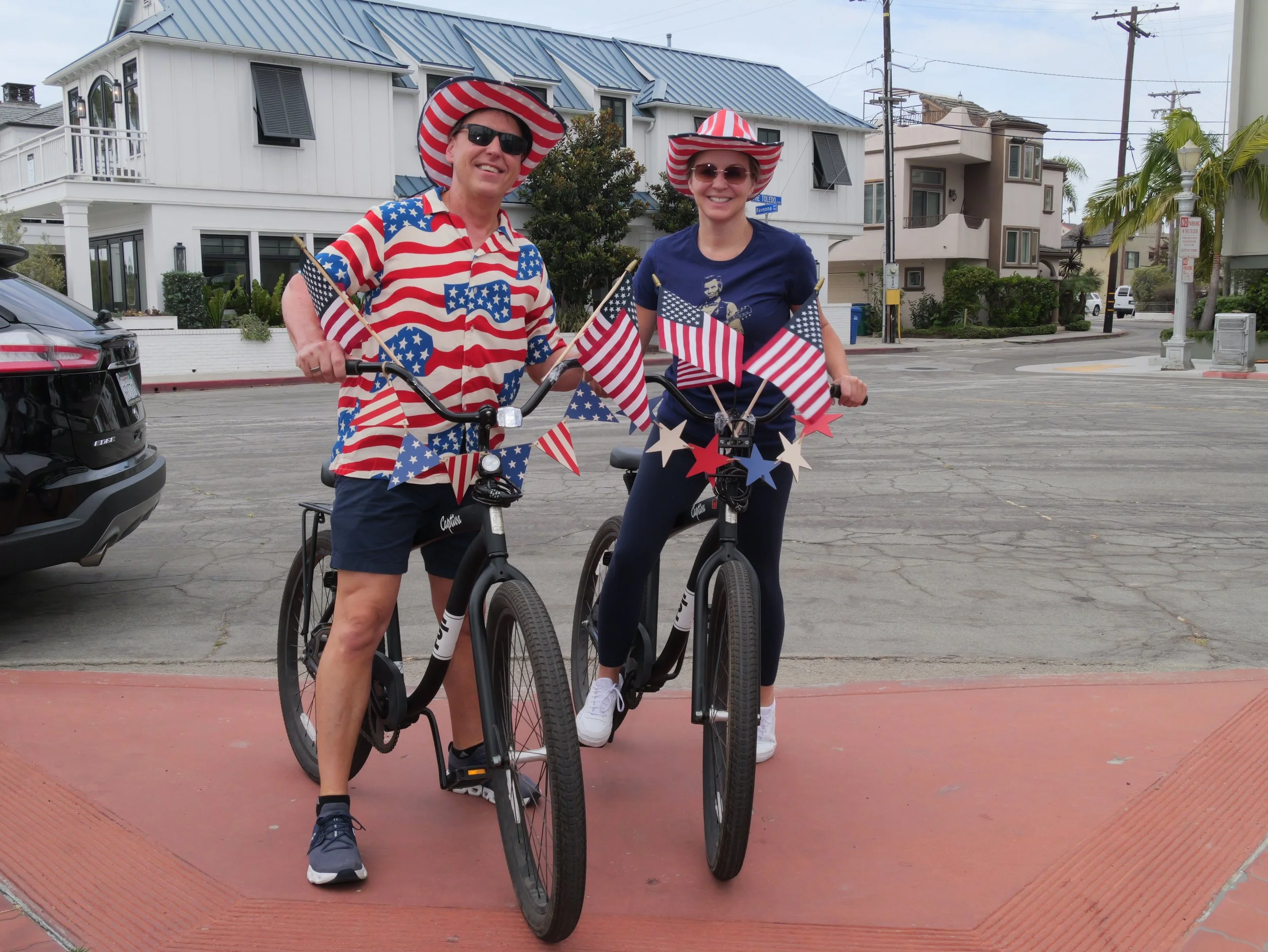 Two people wearing patriotic red, white, and blue clothing and hats, smiling while riding bikes decorated with American flags on a city street.
