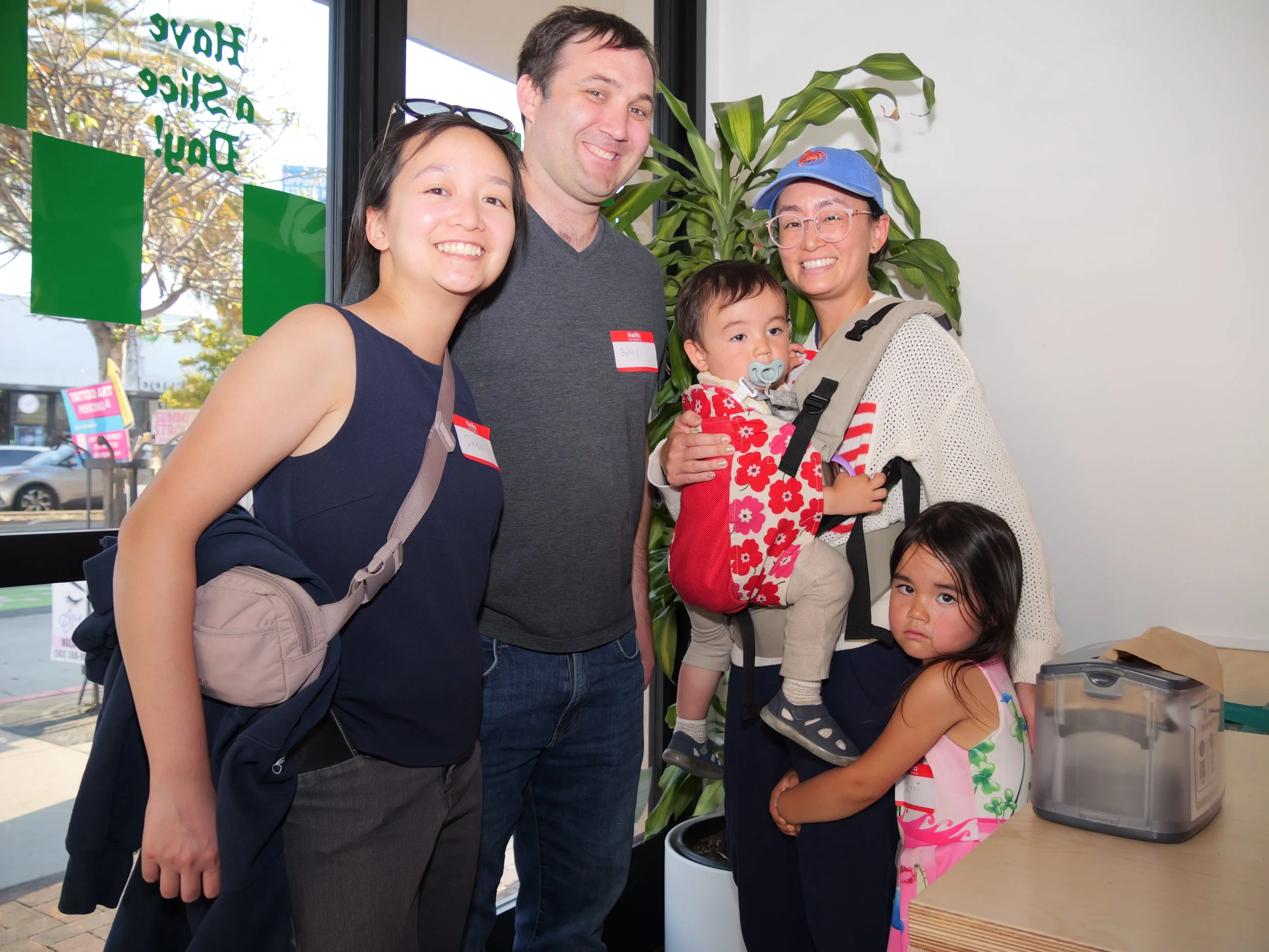 A group of five people, including two women, a man, and two children, standing inside a room near a counter with a food container. They are smiling and looking at the camera, with a large window behind them showing trees and a street outside.