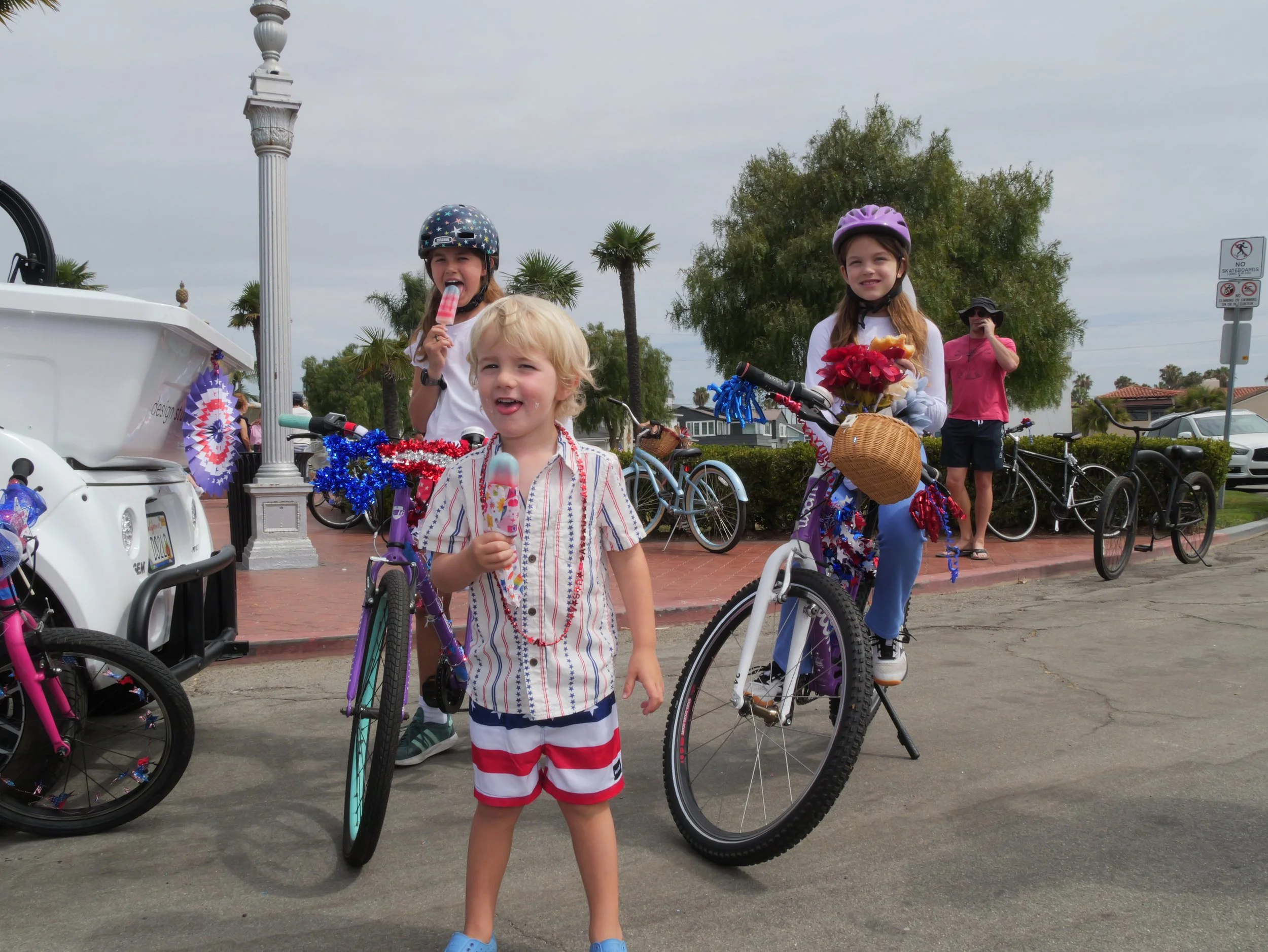 Three children with helmets and bikes, one boy holding a popsicle, in a park-like setting with trees and parked cars in the background.